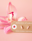 On a pink background is a gold plated of pair of baguette stud earrings with a pink stone in the center.
