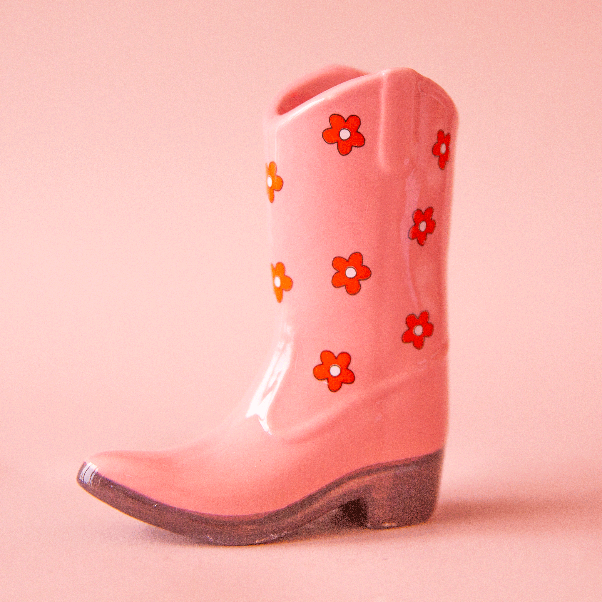 On a pink background is a pink cowboy boot shaped vase with a red and white daisy print on it.