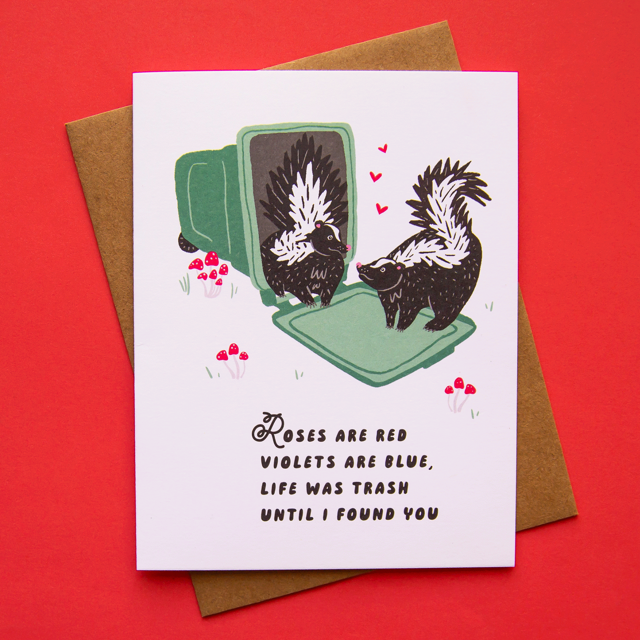 A white card with an illustration of two skunks standing on a tipped over garbage can with red mushrooms around them and pink hearts floating between along with black text on the bottom half that reads, "Roses are red, violets are blue, life was trash until I found you".