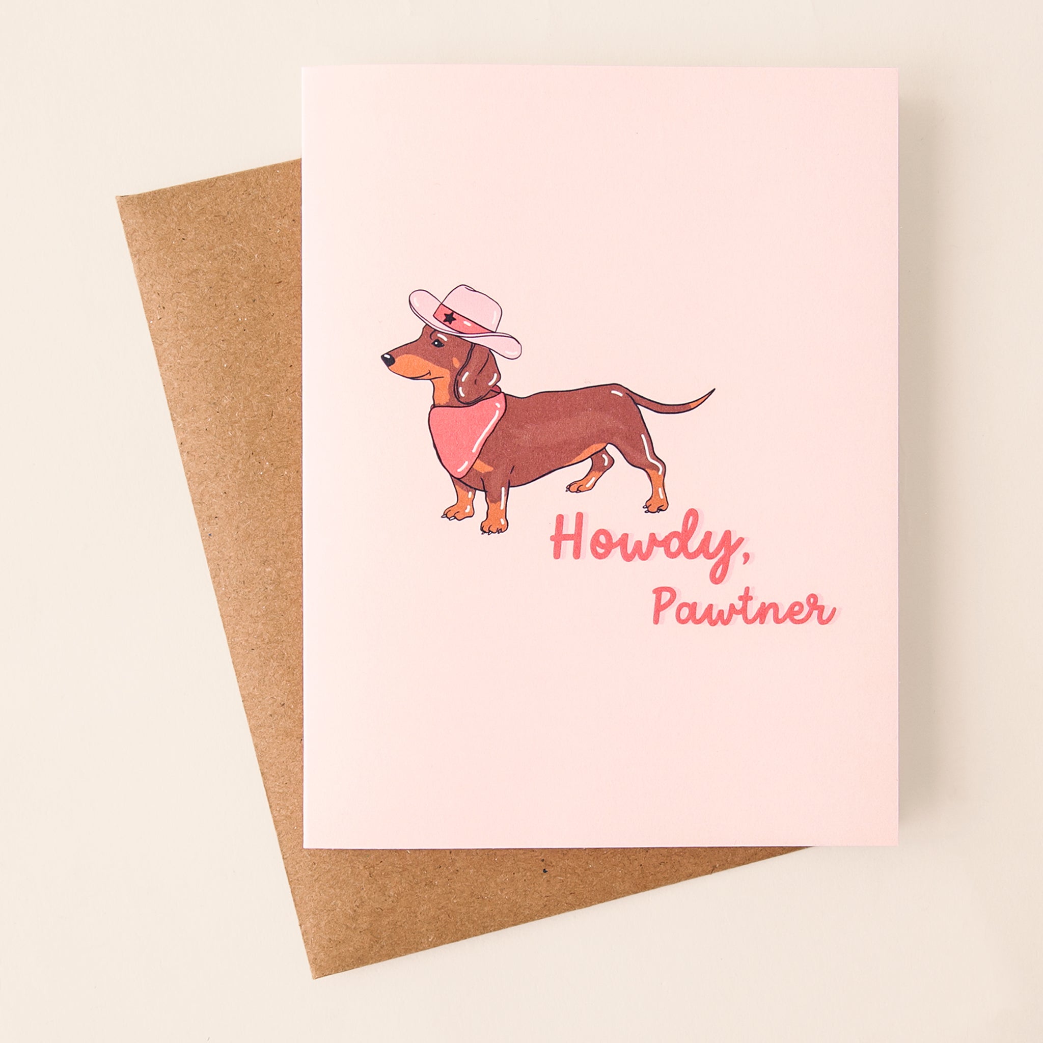 On a tan background is a light pink card with an image of a Weiner dog in a pink bandana and a western cowgirl hat along with pink cursive text to its right that reads, "Howdy Partner".