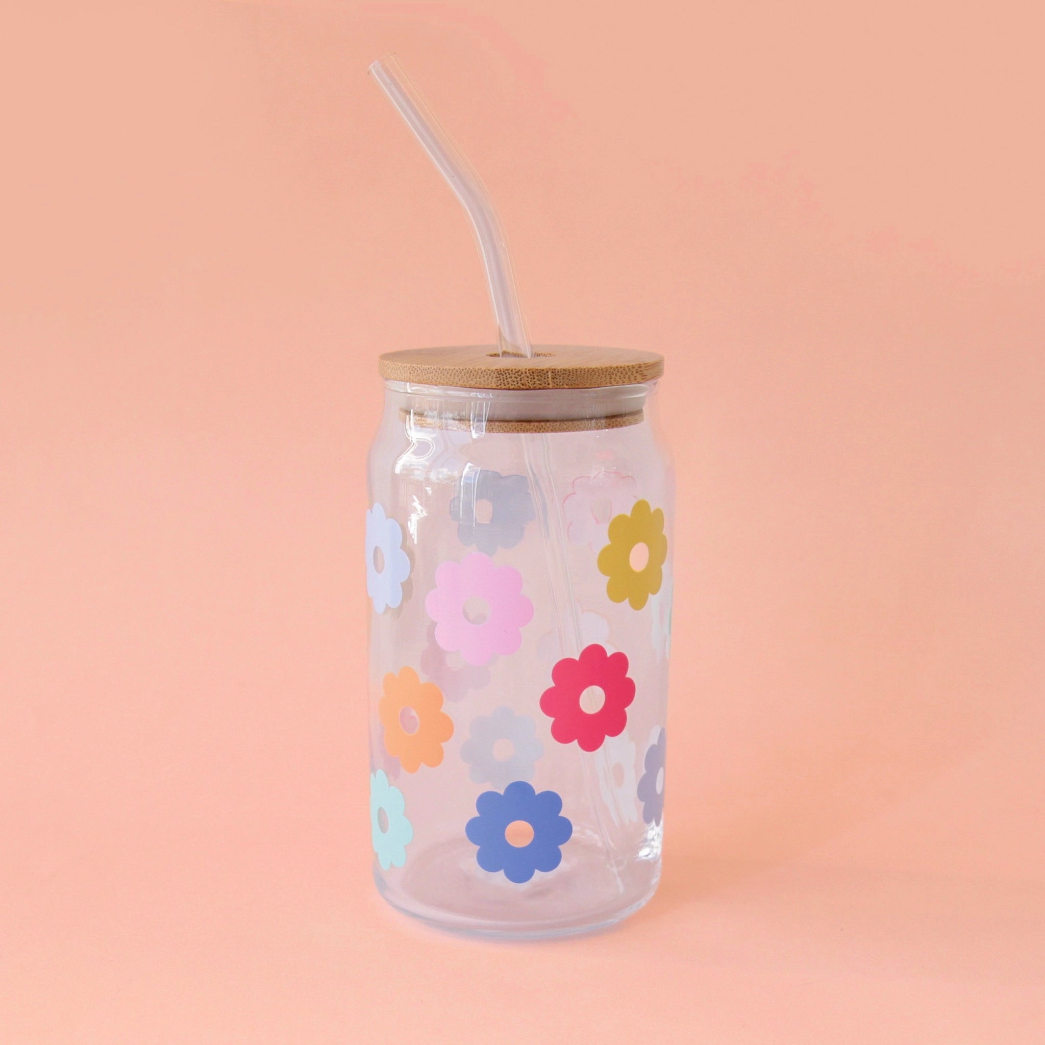 A glass tumbler with multi colored daisies and a wooden lid and a glass straw.