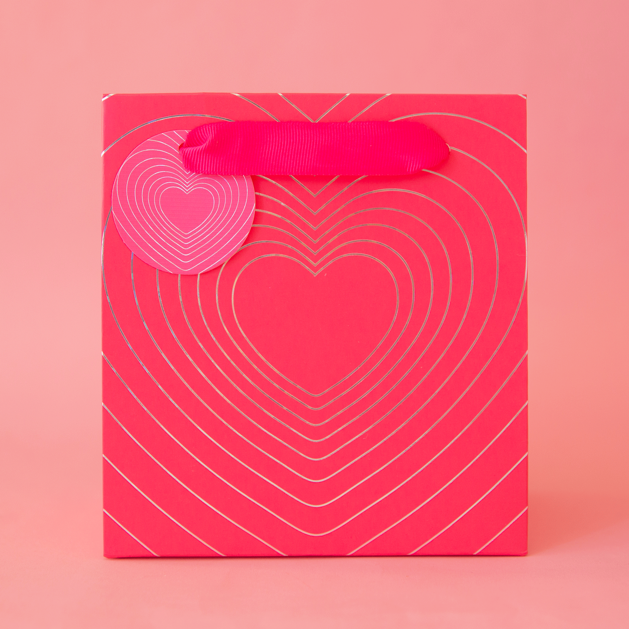 On a pink background is a small gift bag with a radiating heart design and ribbon handles.