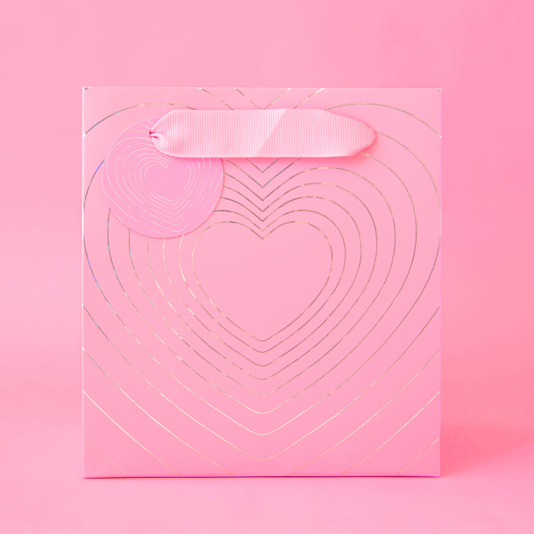 On a light purple background is a purple gift bag with a silver radiating heart design and ribbon handles.