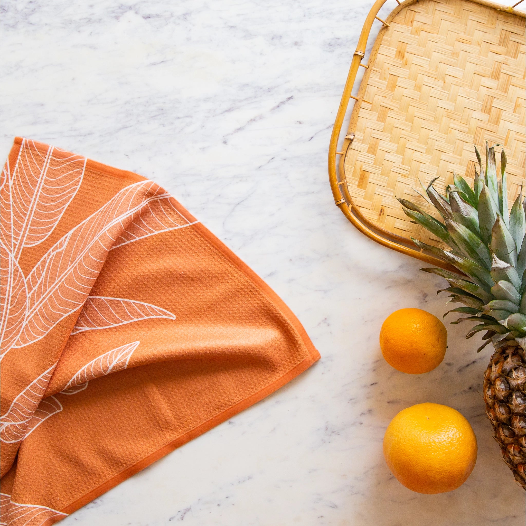 On a marble background is an orange kitchen towel with a white banana leaf design.