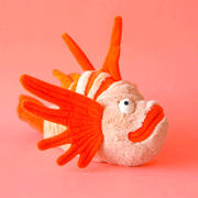 On a coral background is orange lionfish shaped stuffed toy.