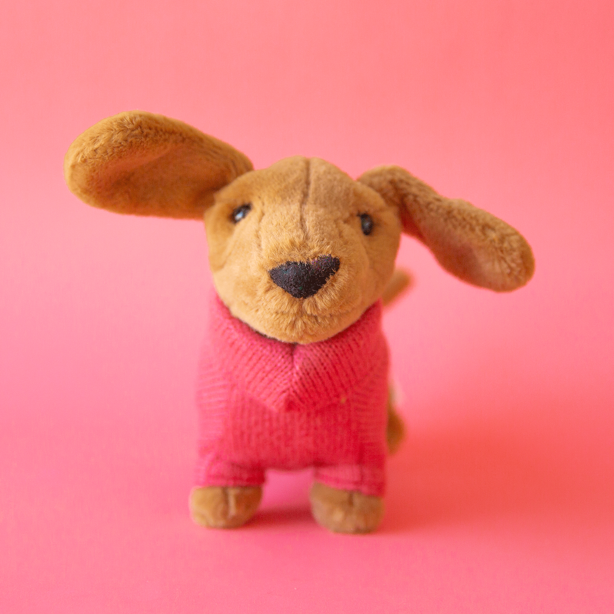 On a pink background is a tan sausage dog stuffed animal wearing a pink sweater.