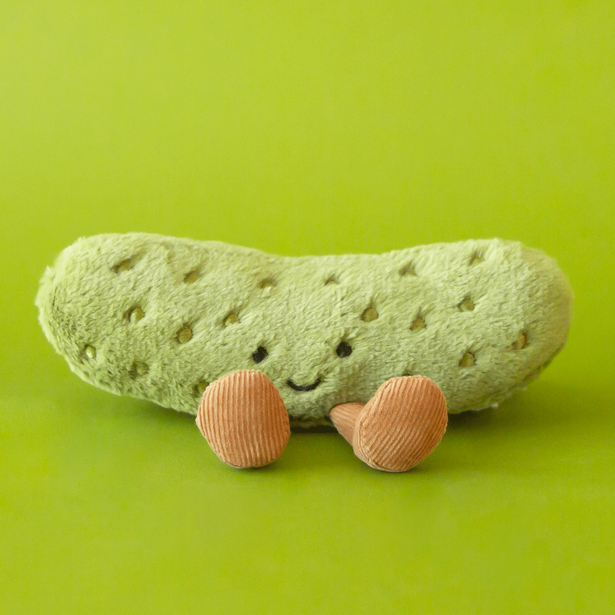 On a green background is a green pickle shaped stuffed toy with a smiling face and brown corduroy legs and feet. 