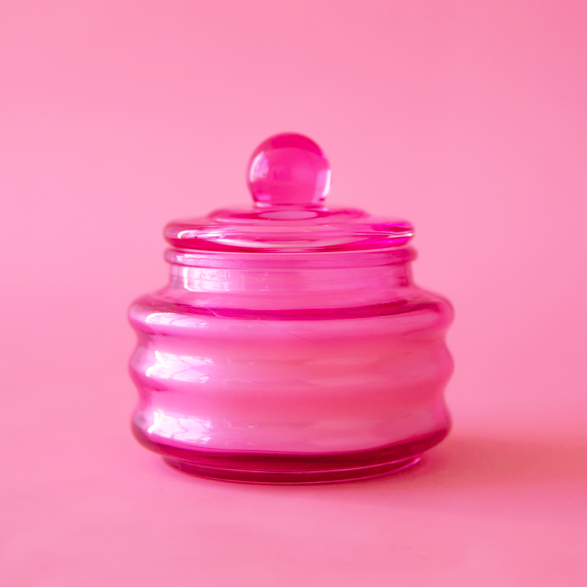 On a pink background is a glass pink jarred candle with a coordinating lid. 
