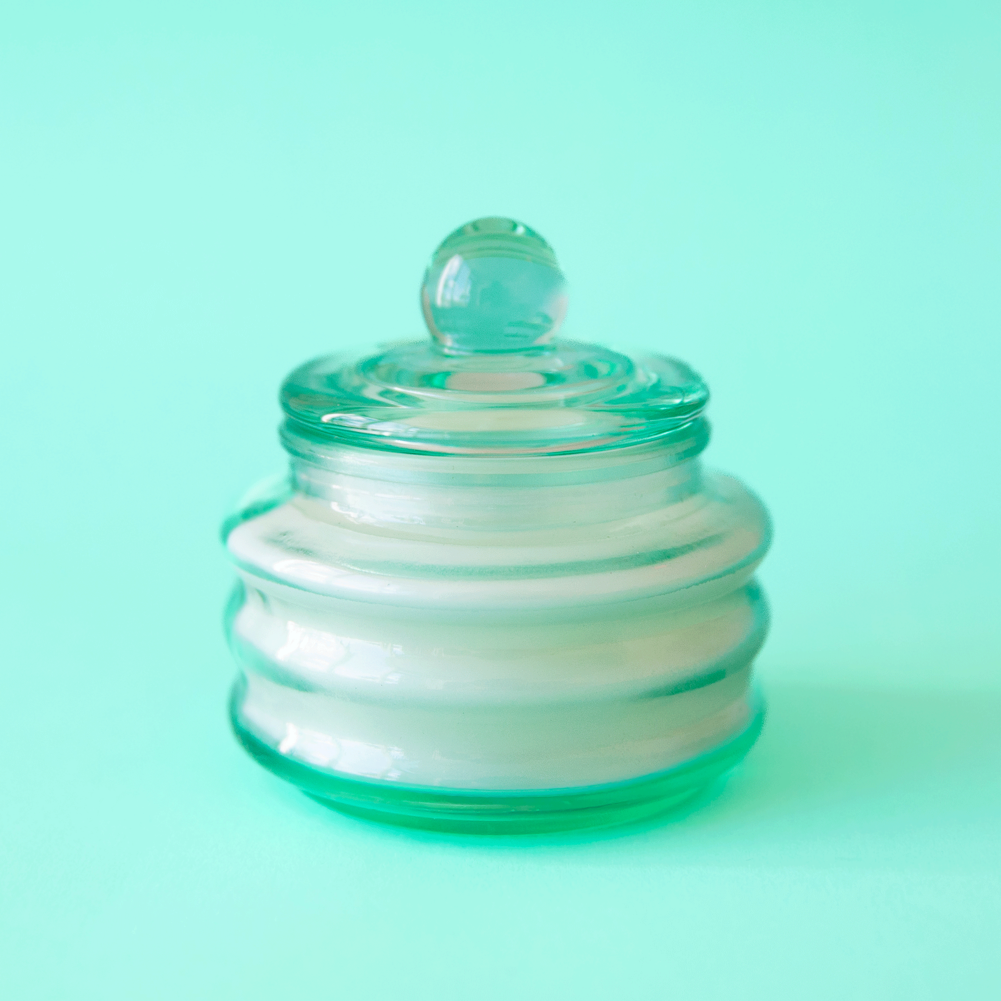 On a turquoise background is a glass jar candle with a lid. 