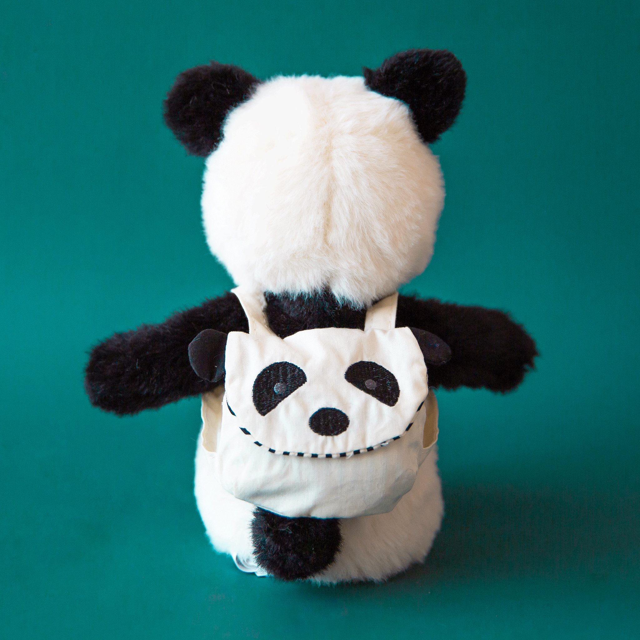 On a teal background is a black and white panda stuffed toy wearing a panda backpack.