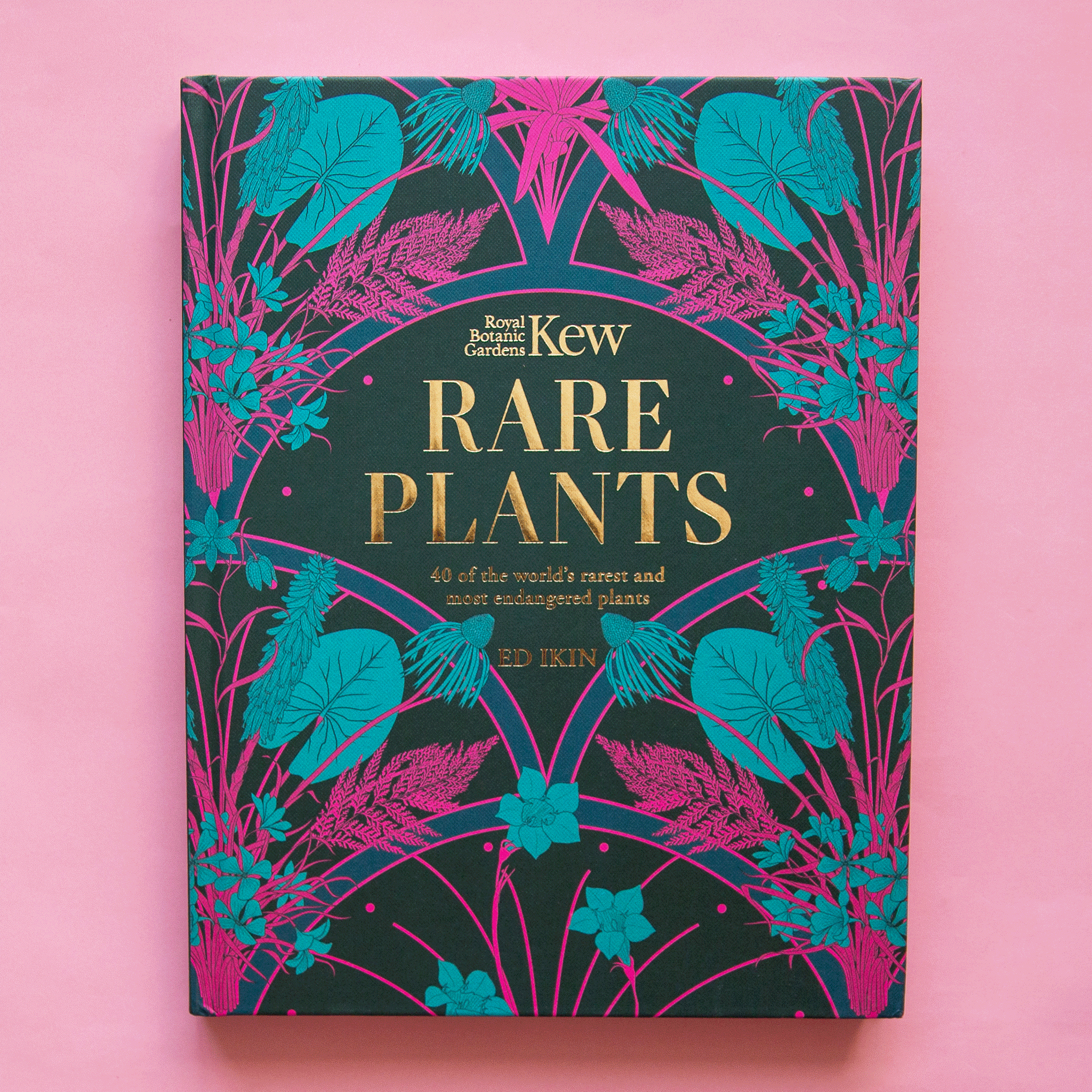 On a pink background is a dark green book cover with a blue and hot pink floral and plant print along with a gold title in the center that reads, "Kew Rare Plants". 
