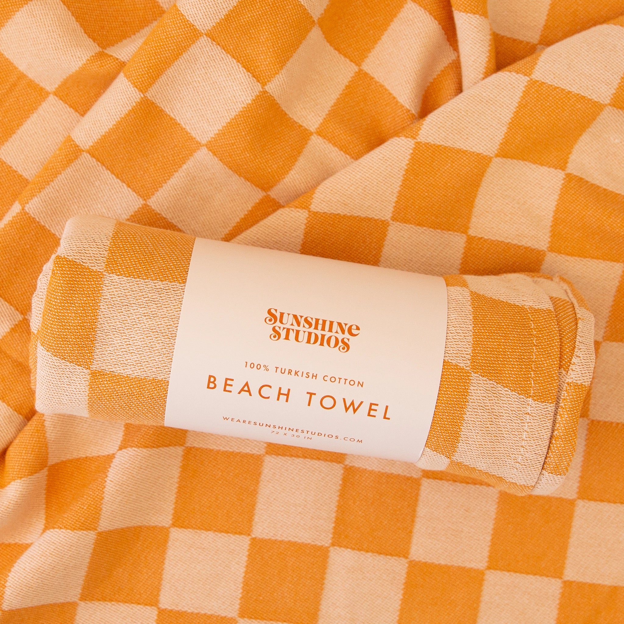 A rolled up beach towel with a orange checkered pattern laying on an opened up towel. 