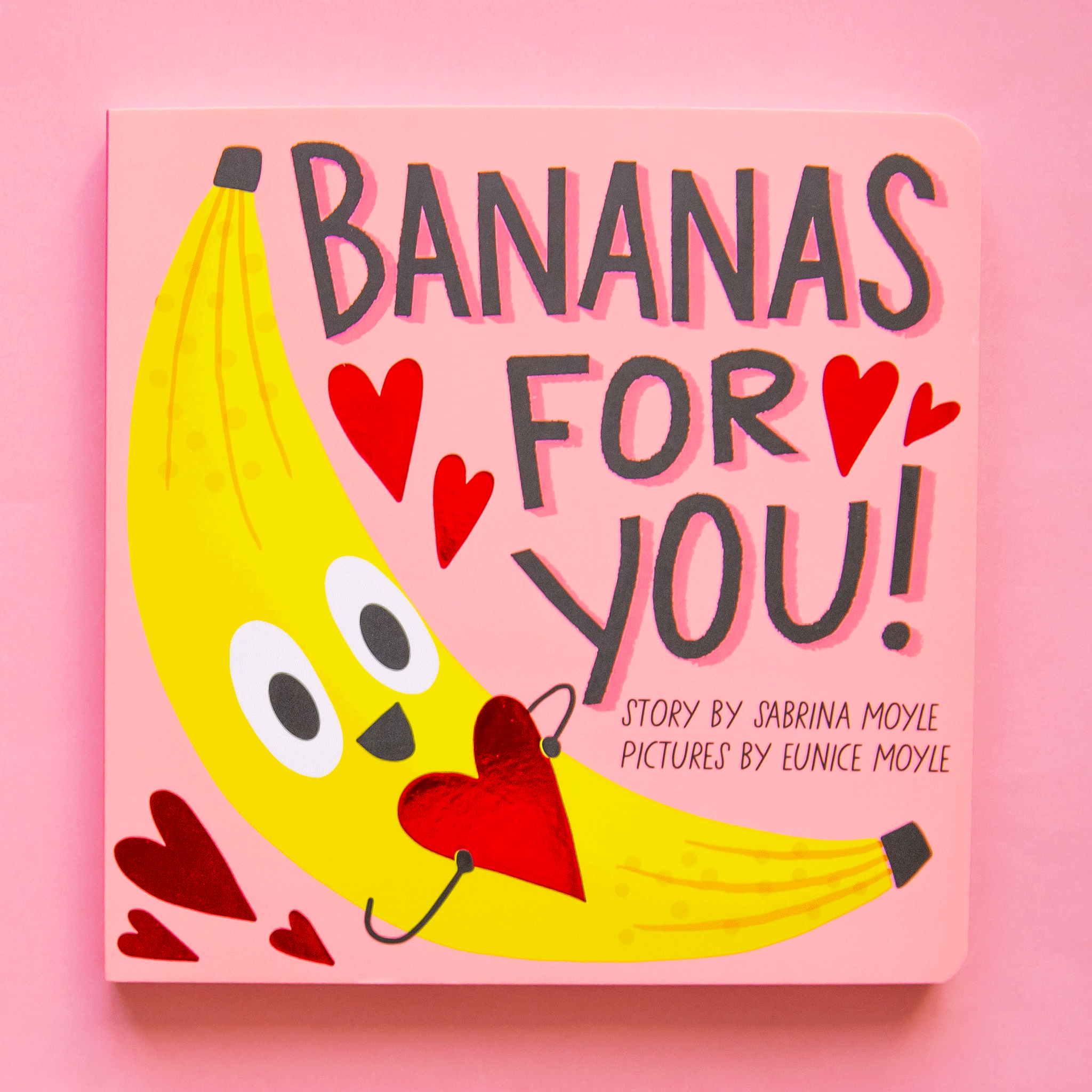 On a pink background is a pink book cover with a banana on the front holding a heart and title that reads, "Bananas For You". 