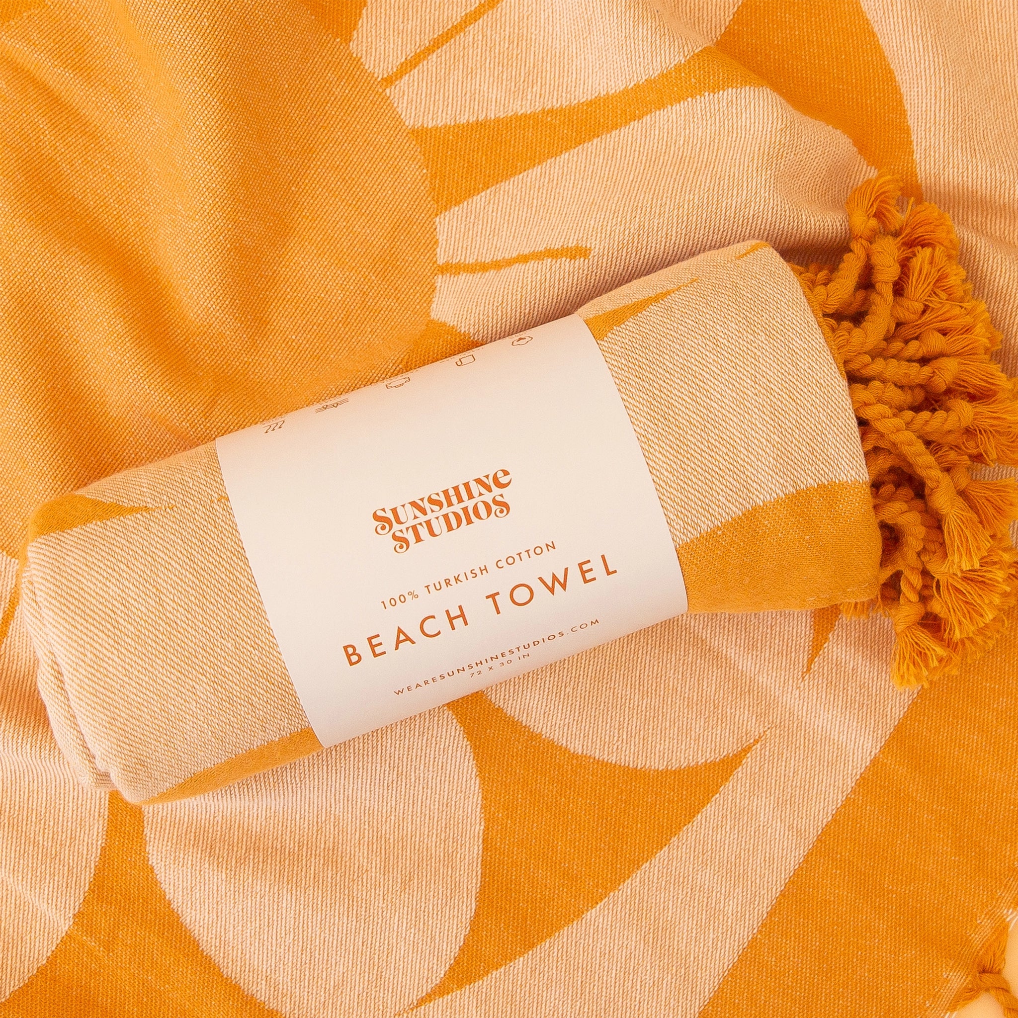 An orange beach towel rolled up with white label that reads, &quot;Sunshine Studios 100% Turkish Cotton Beach Towel&quot; along with a retro flower design and orange string tassels. 