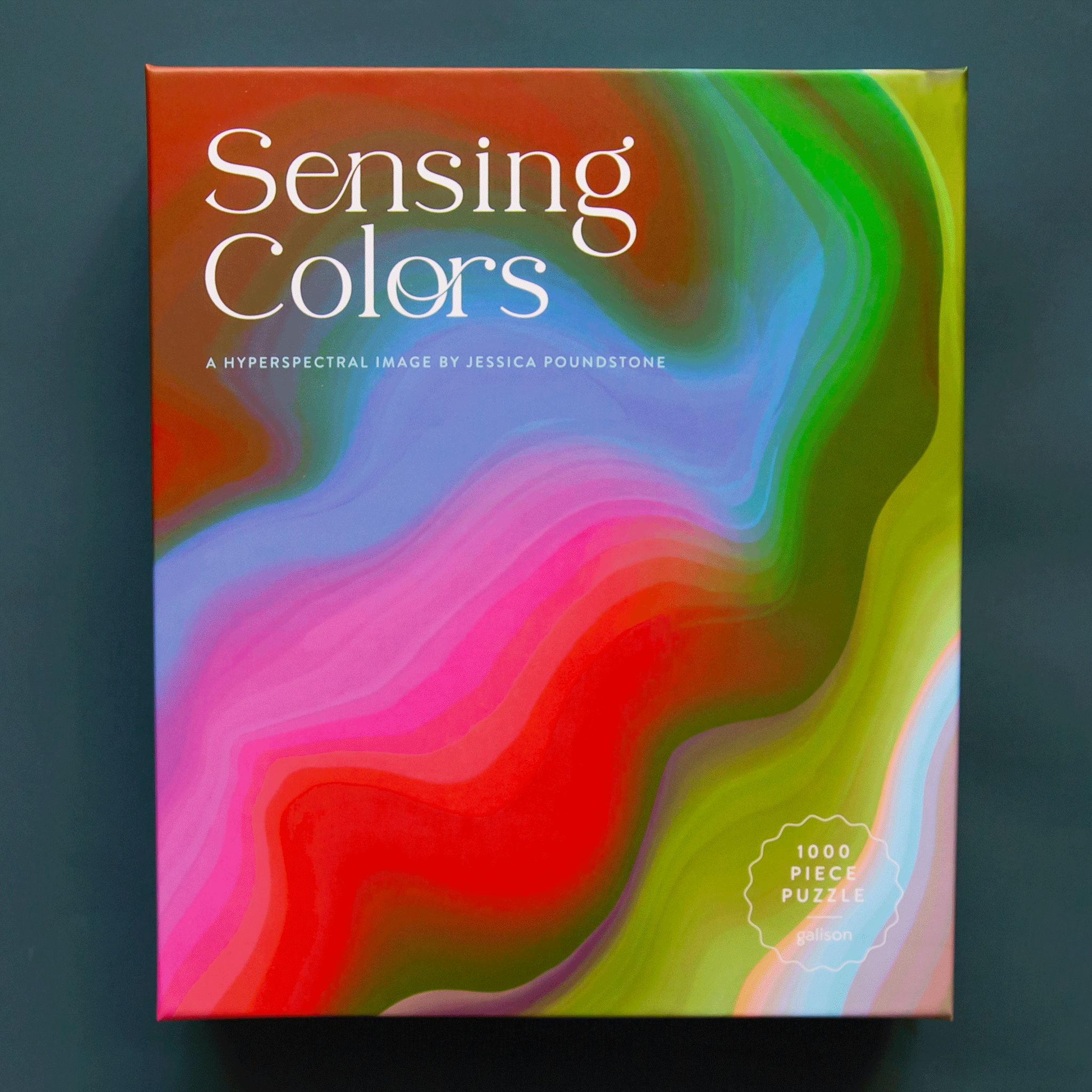 On a dark teal background is a multi colored wavy designed box with a puzzle inside and white text on the front of the box that reads, "Sensing Colors".