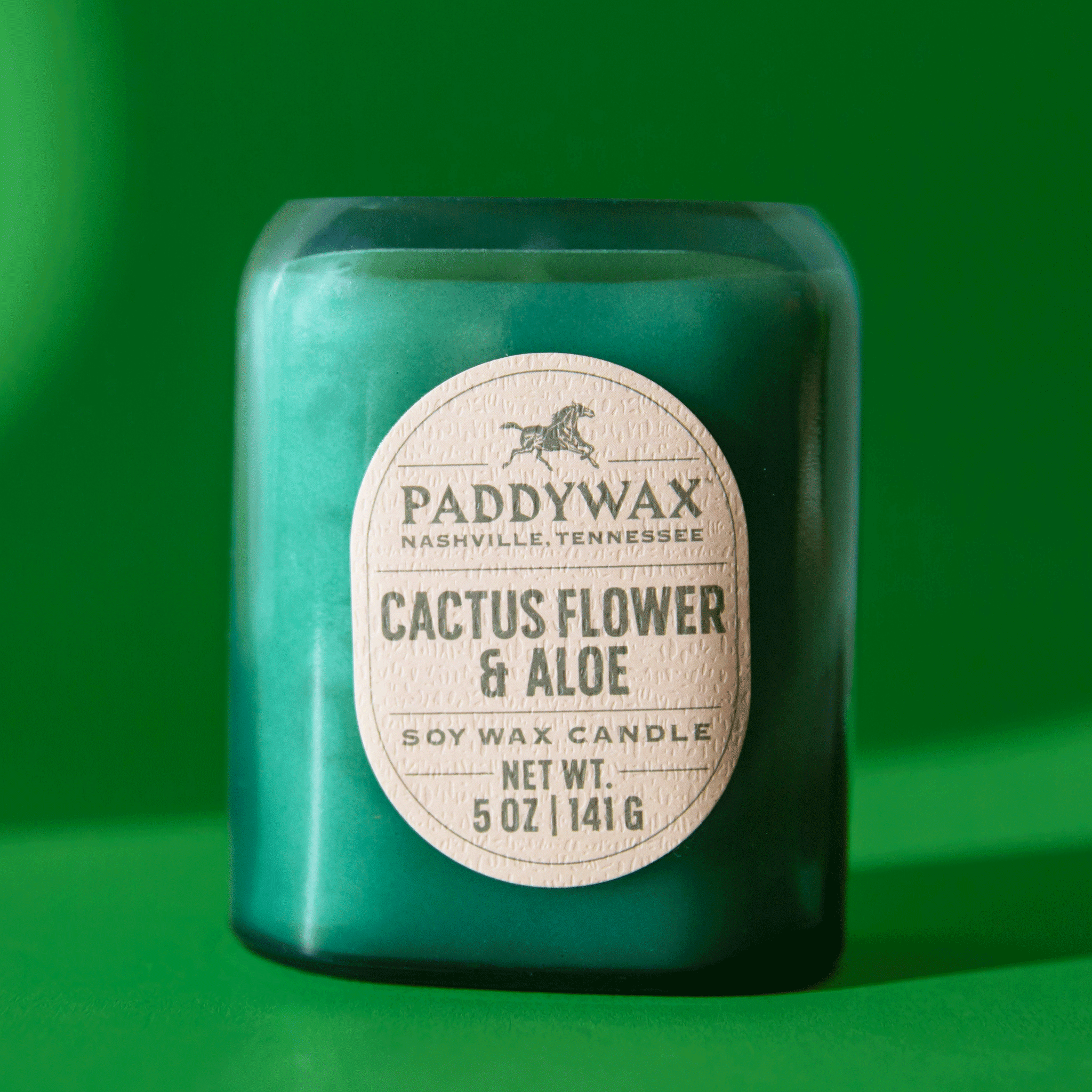 On a green background is a teal glass candle with an oval label that reads, "Paddywax Cactus Flower & Aloe".