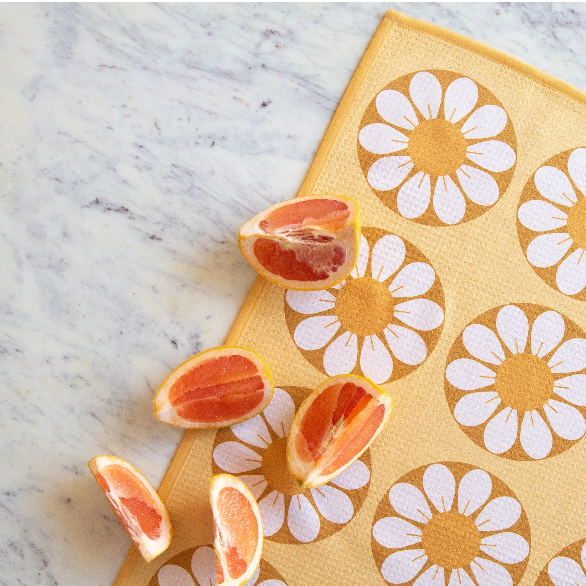 On a marble background is a yellow and white daisy print kitchen towel with a subtle waffle texture staged with blood orange slices. 