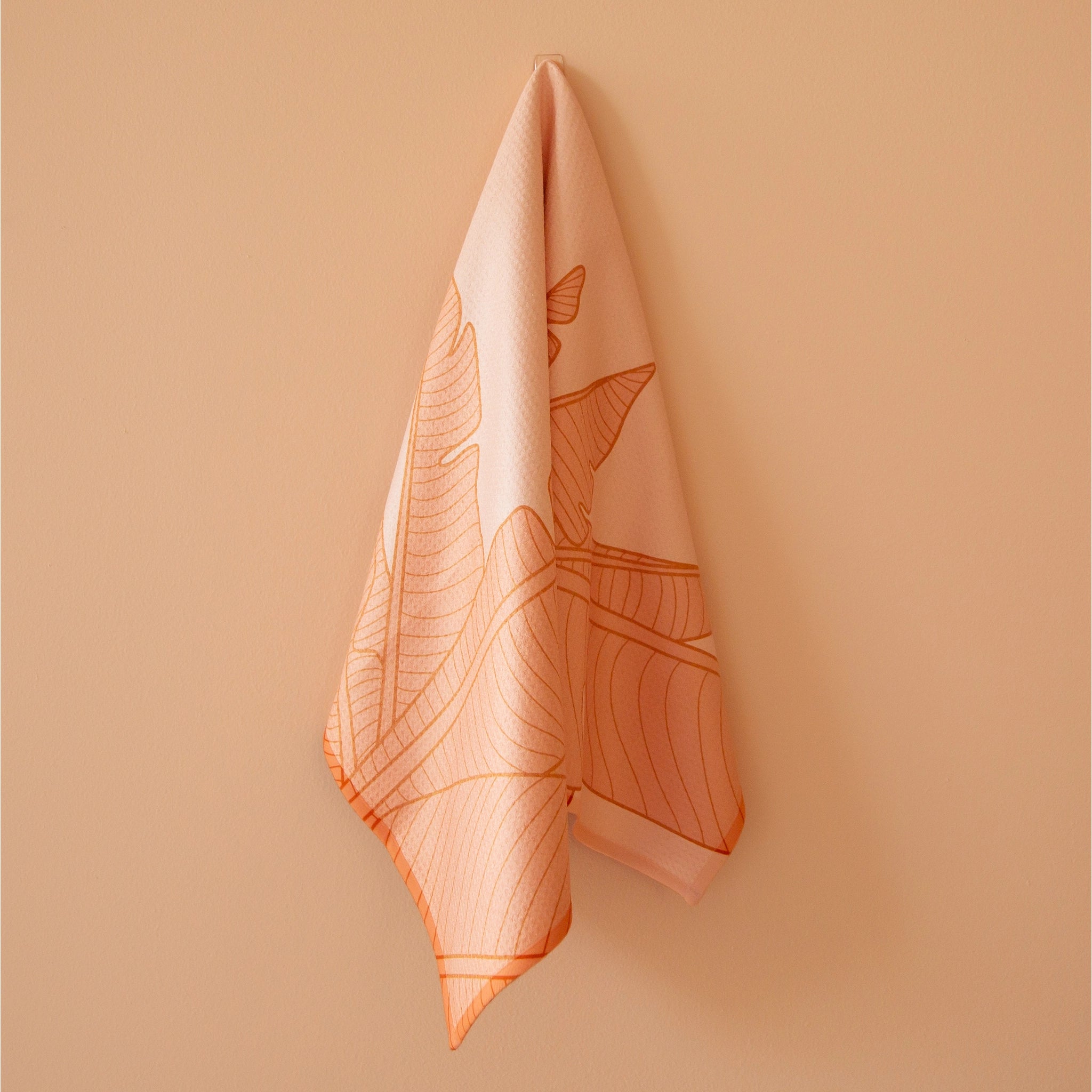 On an orange background is a salmon pink kitchen towel with a bird of paradise pattern. 