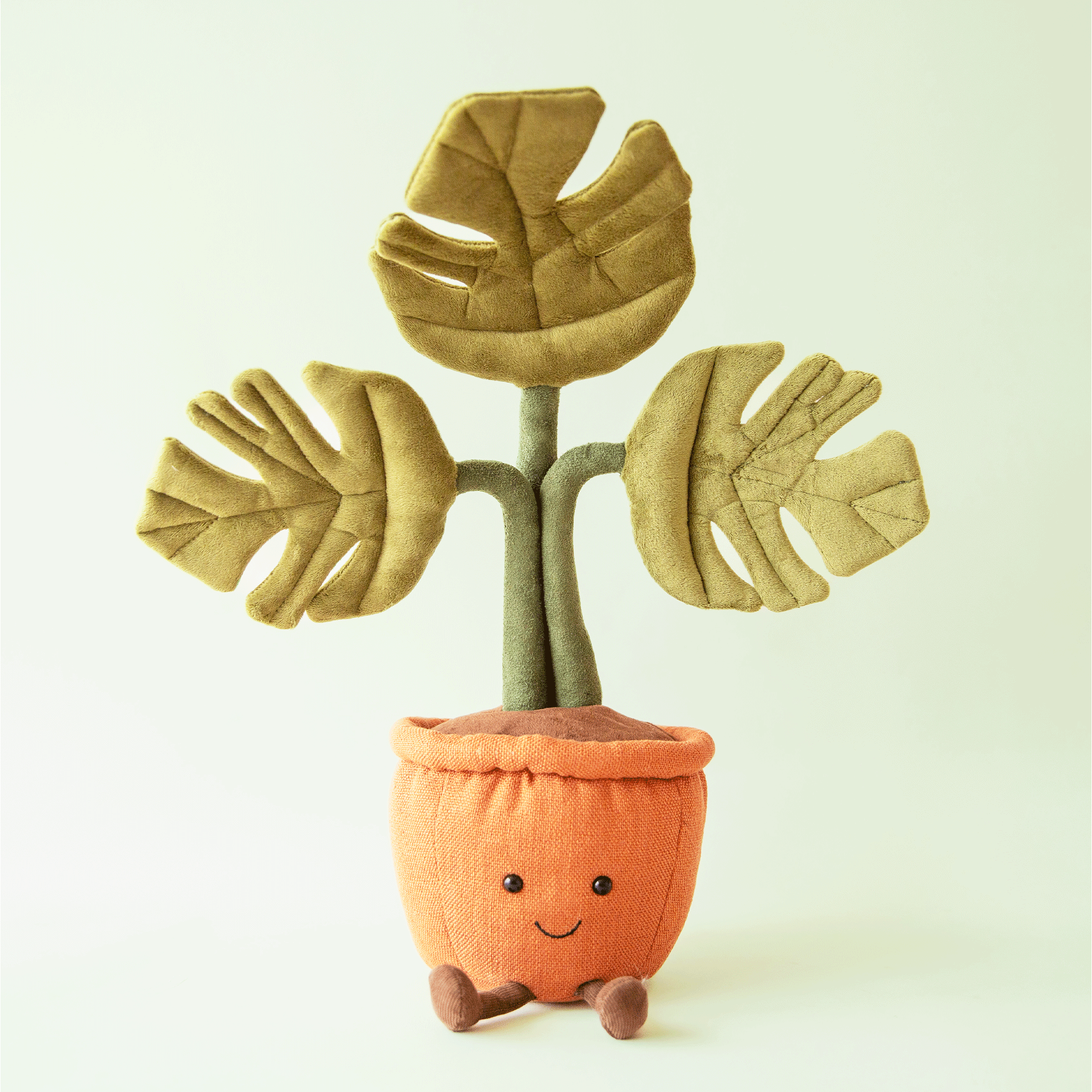 On a mint background is a stuffed toy shaped as a potted monstera plant with a smiling face and three leaves. 