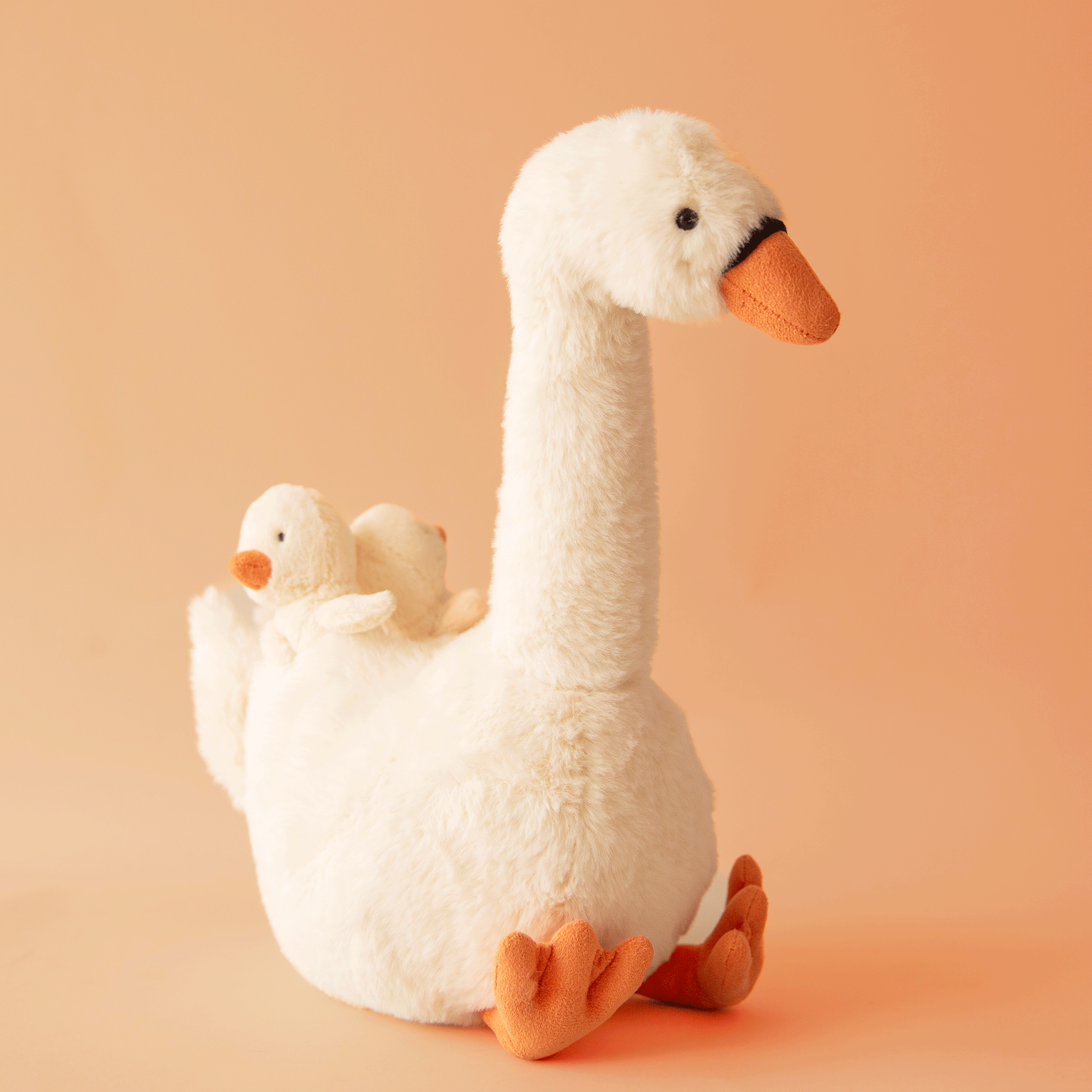 On a orange background is a swan shaped stuffed animal toy with two baby swans on her back.