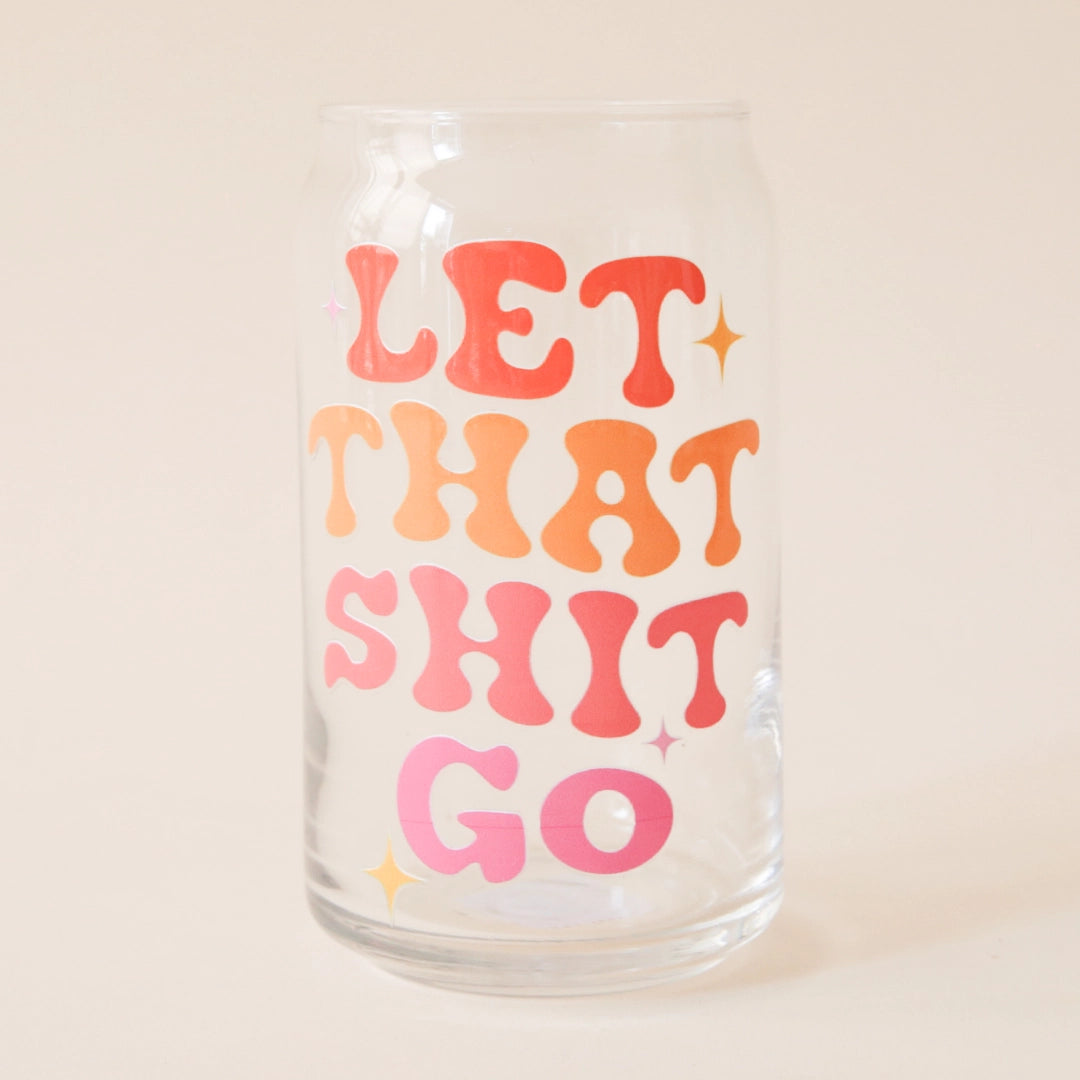 On a cream background is a clear drinking glass with groovy text down the front that reads, &quot;Let That Shit Go&quot; in red, orange and pink letters. 