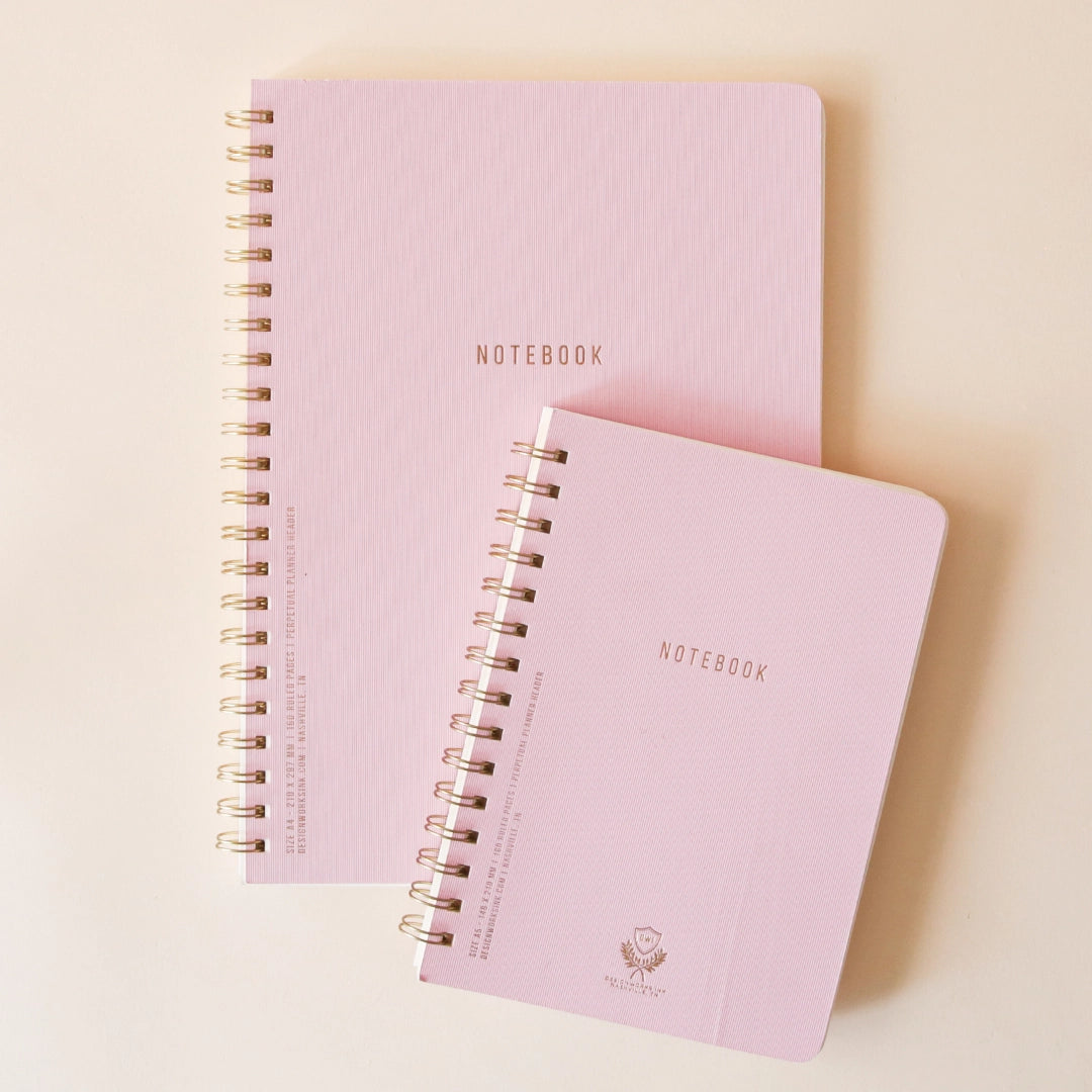 On a beige background is two different sized light purple spiral notebook with small letters on the front that read, "Notebook". 