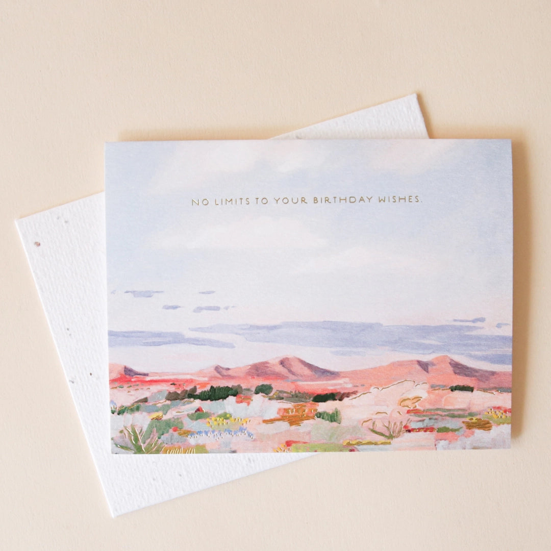 On a cream background is a greeting card with a desert, mountainous landscape illustration with earth tones along with text across the top that reads, "No Limits To Your Birthday Wishes".  
