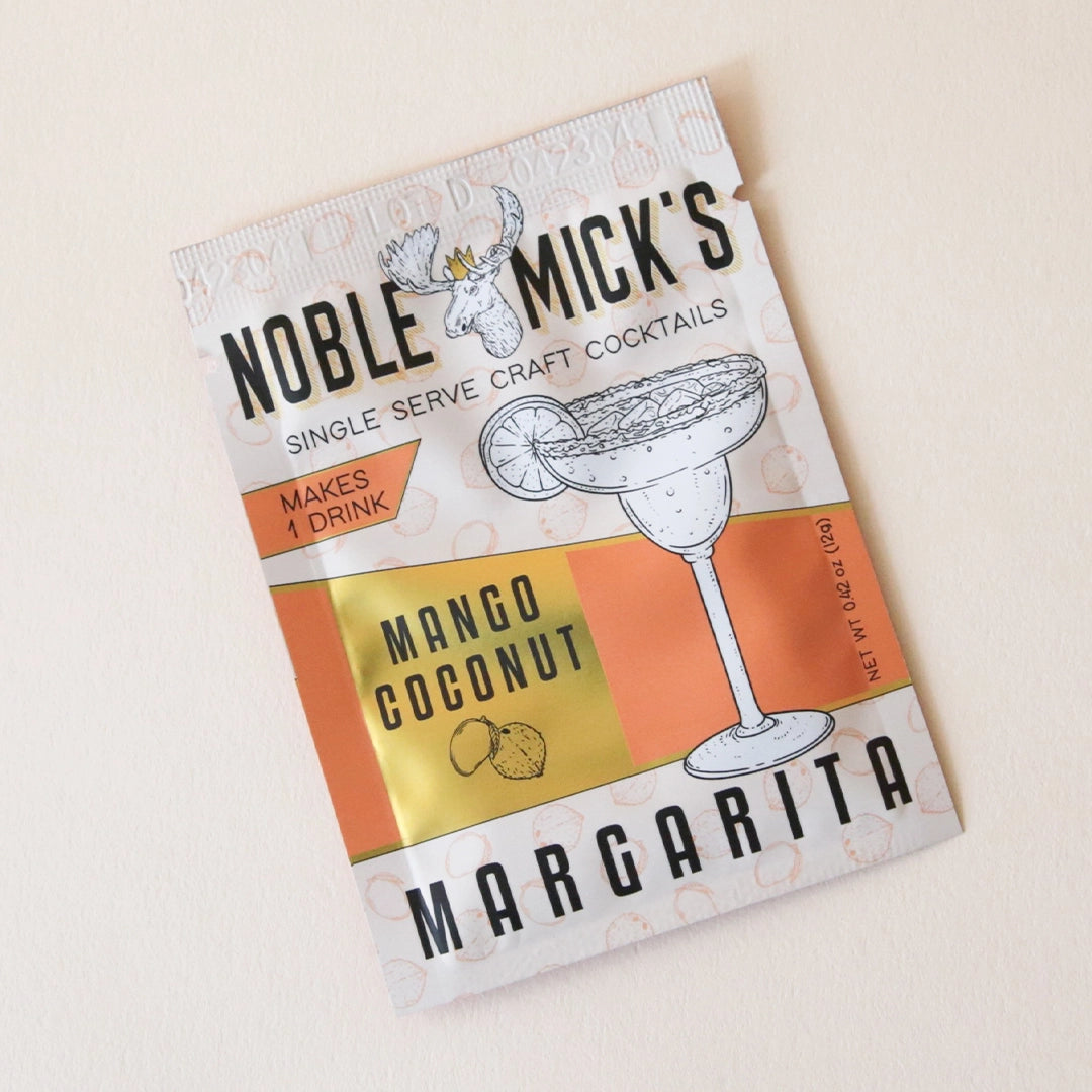 A cream and orange packet of cocktail mix that reads, &quot;Noble Mick&#39;s Single Serve Craft Cocktails Mango Coconut Margarita&quot; in black letters photographed next to a slice of mango.