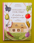 On a green background is a book cover with an illustration of a home with a family in the window and vegetables around the edges and text at the top that reads, "Cocinando On Cook Street A collection of mi familia's recipes". 