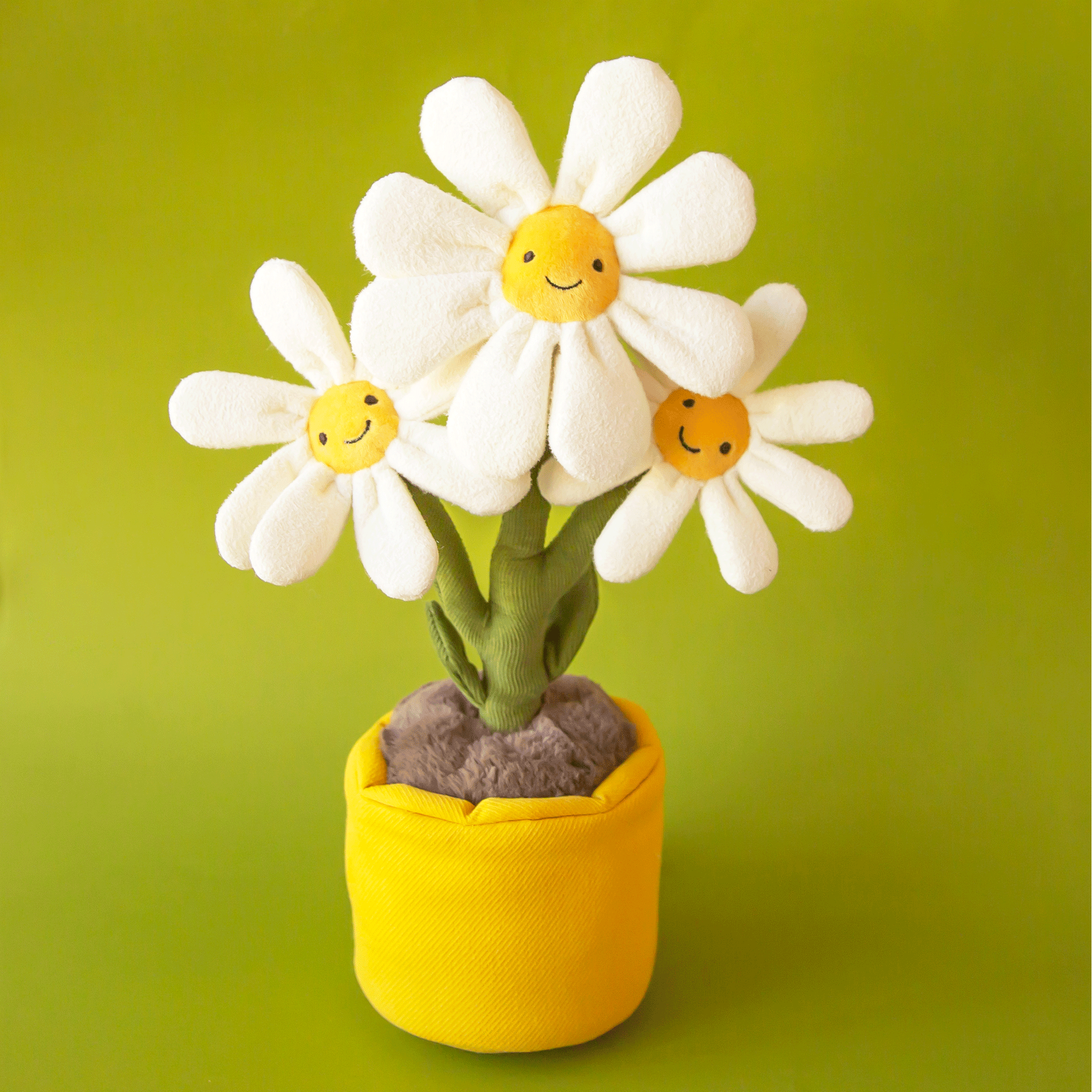 On a green background is a stuffed toy in the shape of three smiling face daisies in a stuffed toy pot.