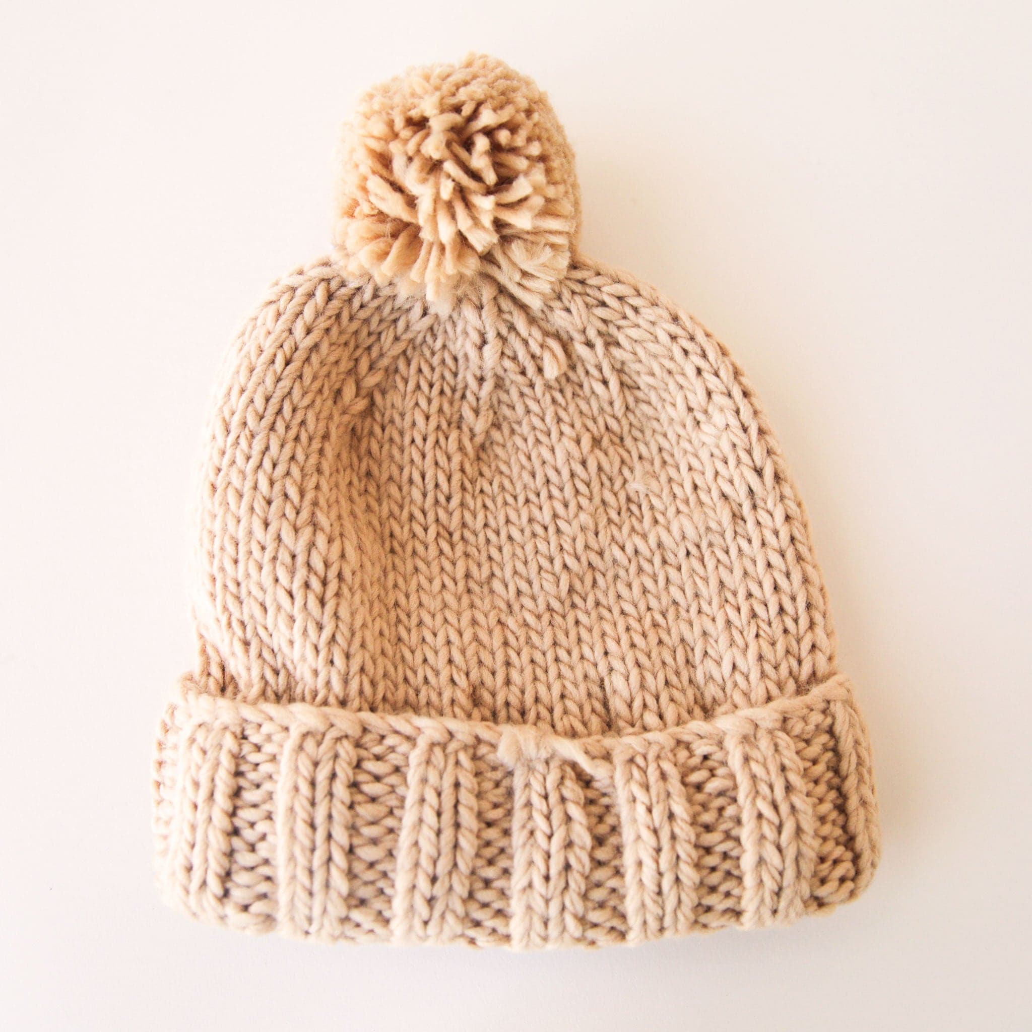 On a white background is a tan knit beanie with a pom pom on top.