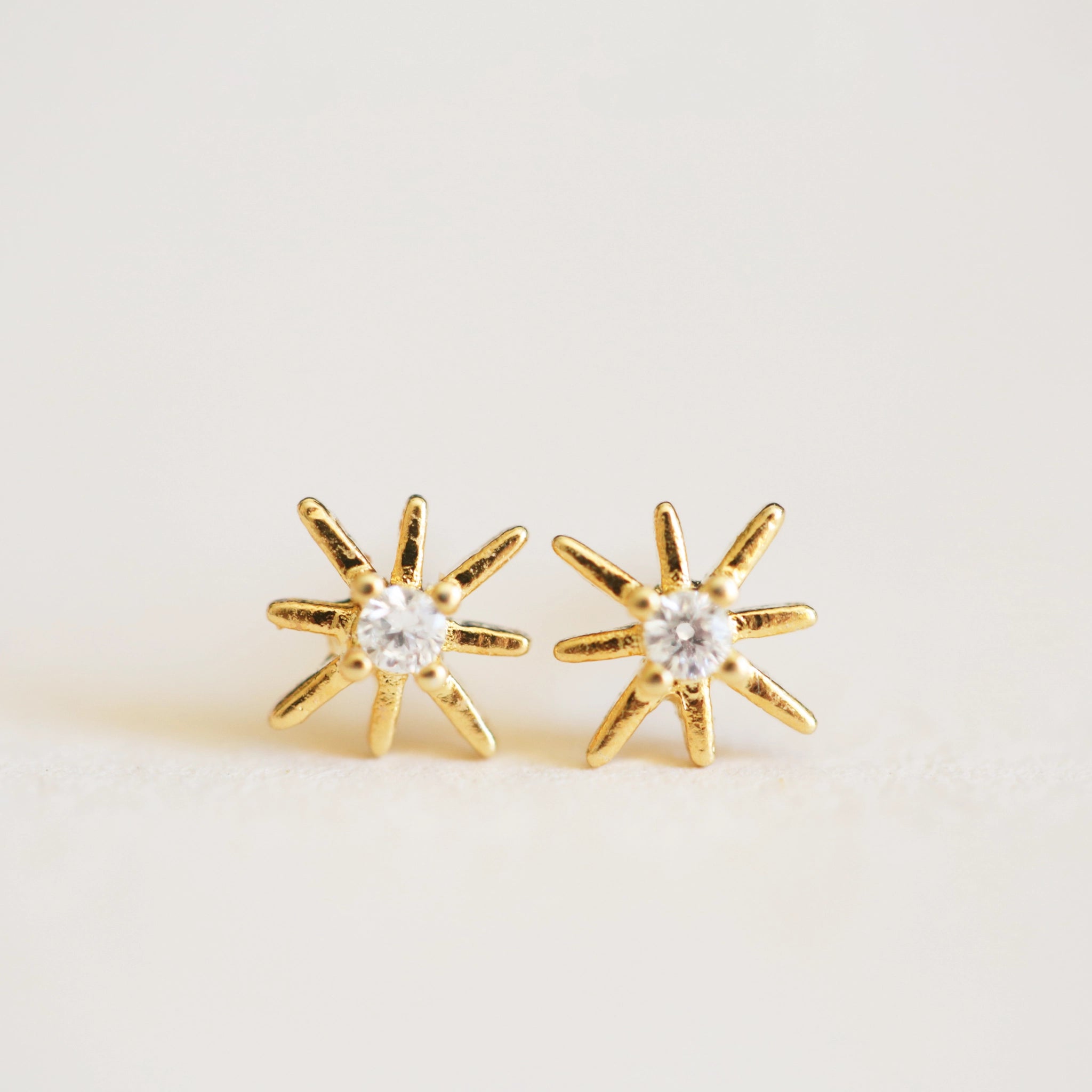 On an ivory background is a pair of dainty gold star earrings with a CZ stone in the center.