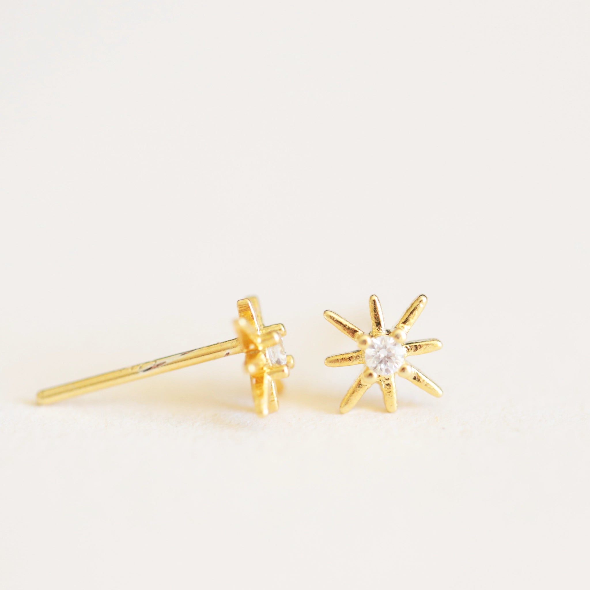 On an ivory background is a pair of dainty gold star earrings with a CZ stone in the center