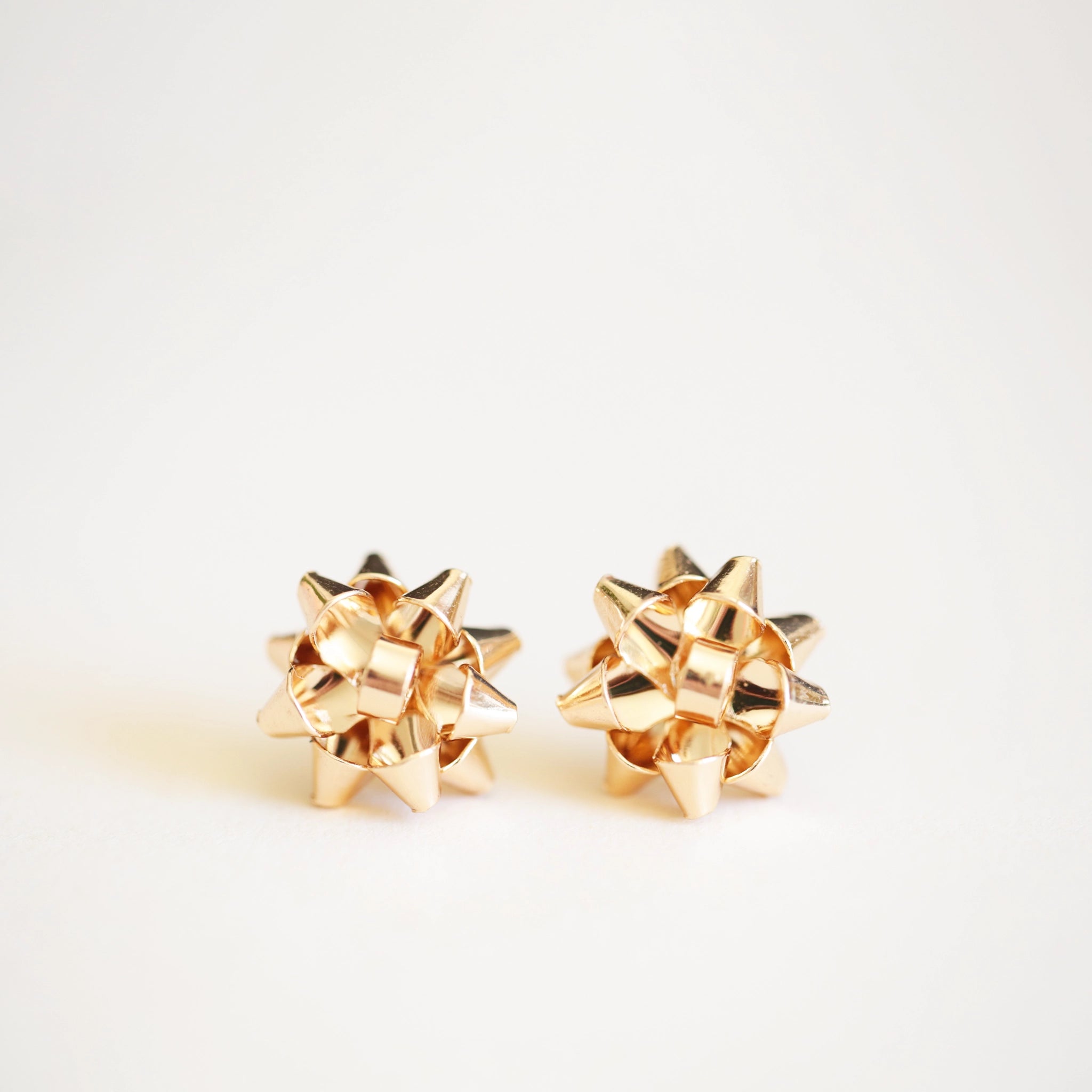 On an ivory background is a pair of gold earrings in the shape of holiday bows.
