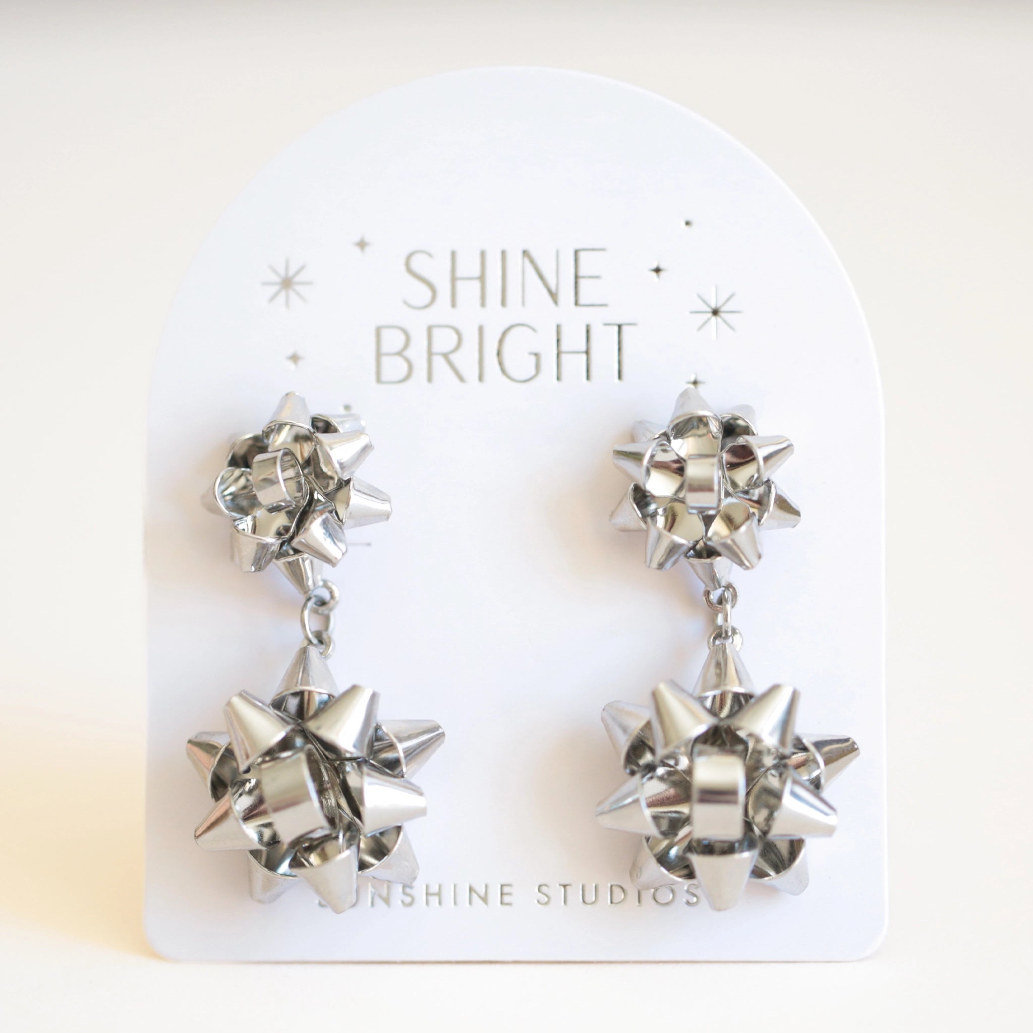 On an ivory background is a pair of silver holiday bow shaped earrings that have a smaller gold bow and a dangling larger bow attached to the bottom. The packaging reads, &quot;Shine Bright Sunshine Studios&quot;.