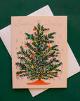 A cream colored card sits against a dark green background. On the front of the card there is a drawing of a dark green Christmas tree. There are yellow and red Christmas lights up and down the tree. There are green, red and pink ornaments spread out all over the tree. Silver tinsel is hanging on the end of each tree branch. At the bottom is a red tree skirt. At the top is a sliver star. Above the tree in silver script it reads ‘season's greetings.’ The card is accompanied by a white envelope.