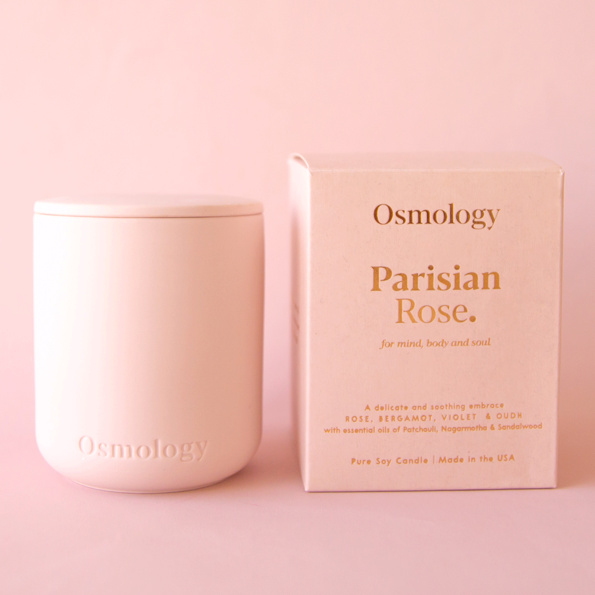 On a pink background is a pink clay jarred candle with a lid and the box that it comes in on the side that reads, "Parisian Rose for mind body and soul.".