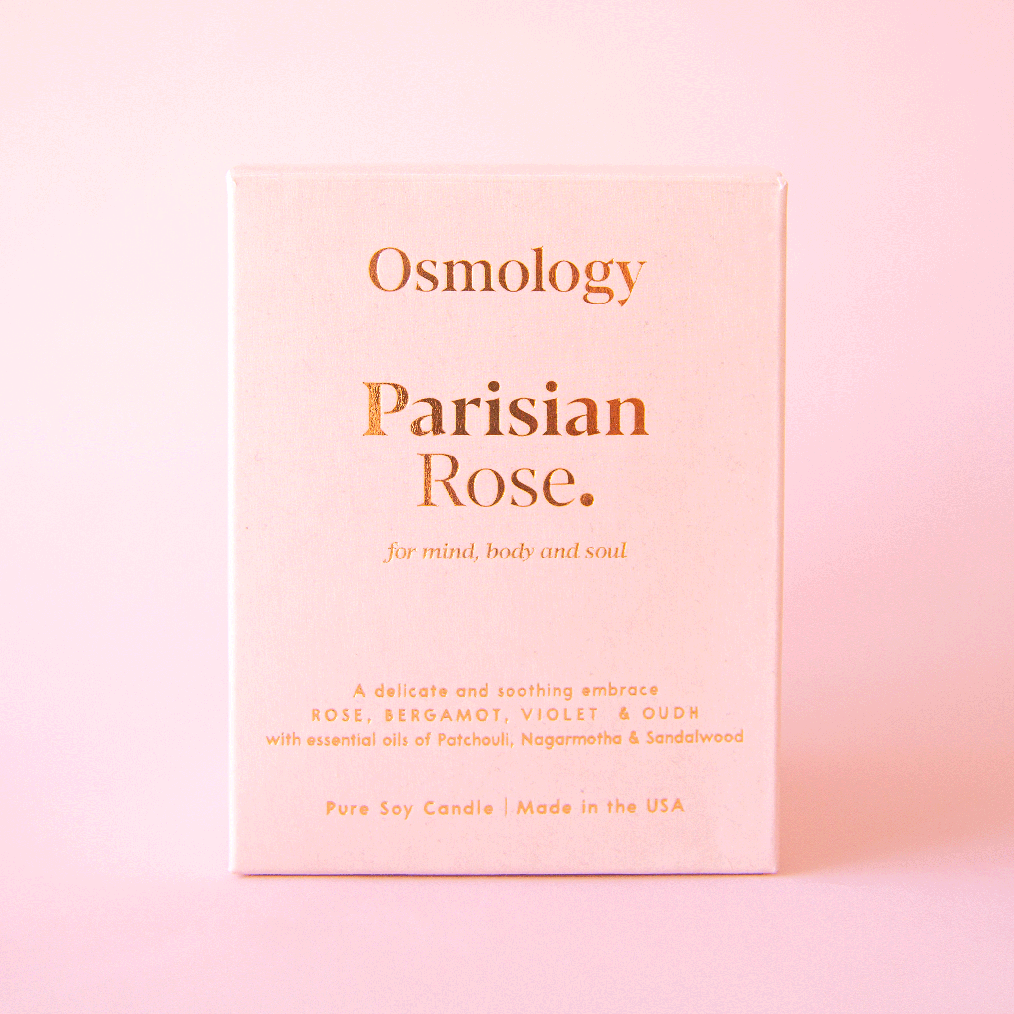 On a pink background is a pink clay jarred candle with a lid and the box that it comes in on the side that reads, &quot;Parisian Rose for mind body and soul.&quot;.