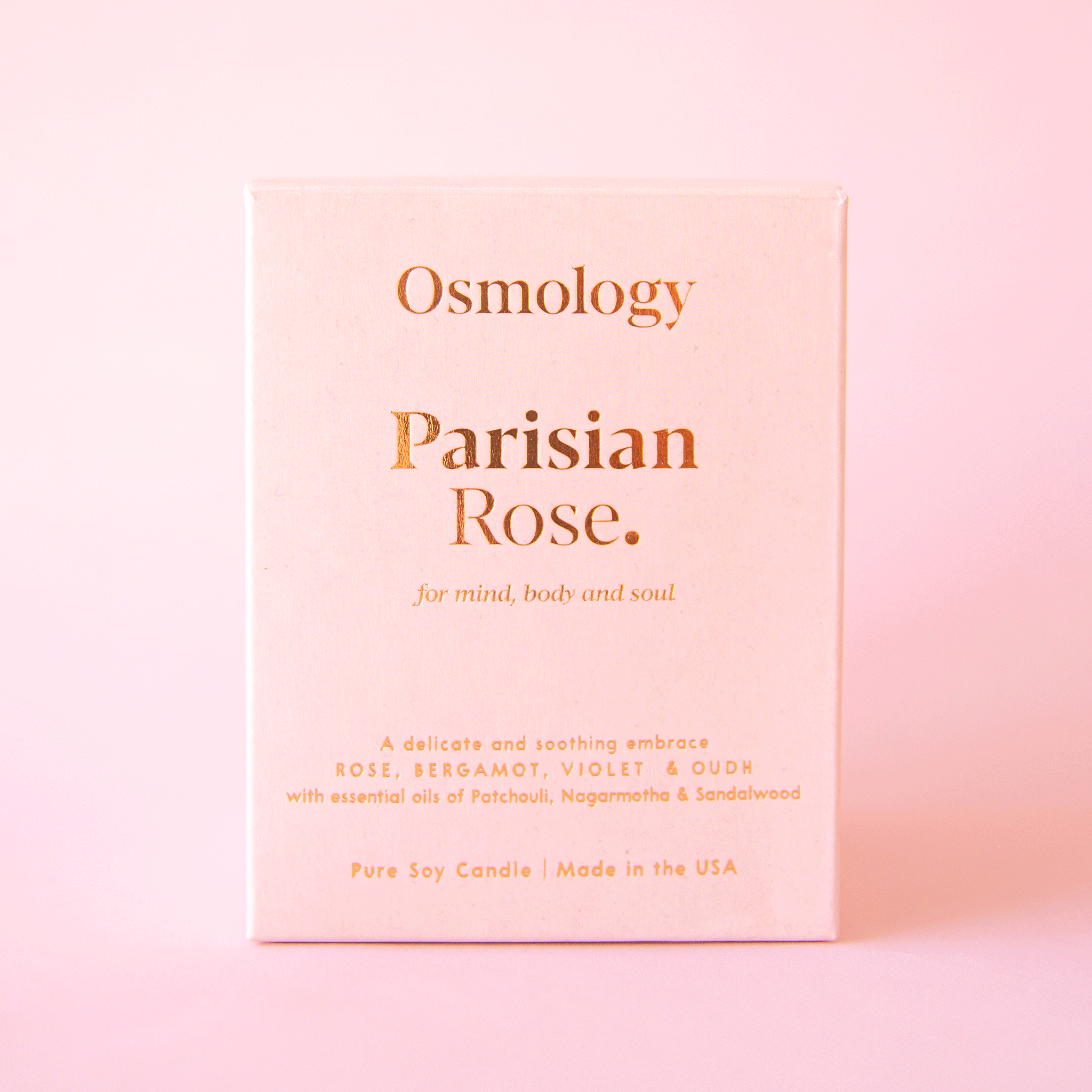 On a pink background is a pink clay jarred candle with a lid and the box that it comes in on the side that reads, "Parisian Rose for mind body and soul.".