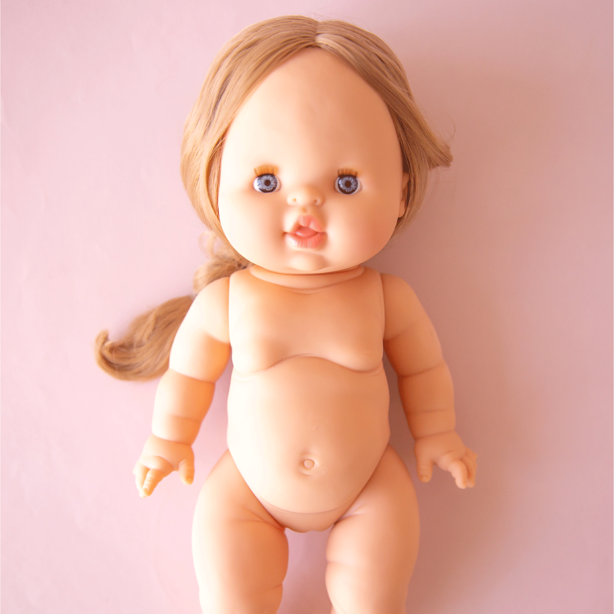 On a pink background is a baby doll with blonde braided hair and blue eyes. 