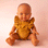 An African American baby girl doll wearing a pink romper jumpsuit in front of a pink background. .