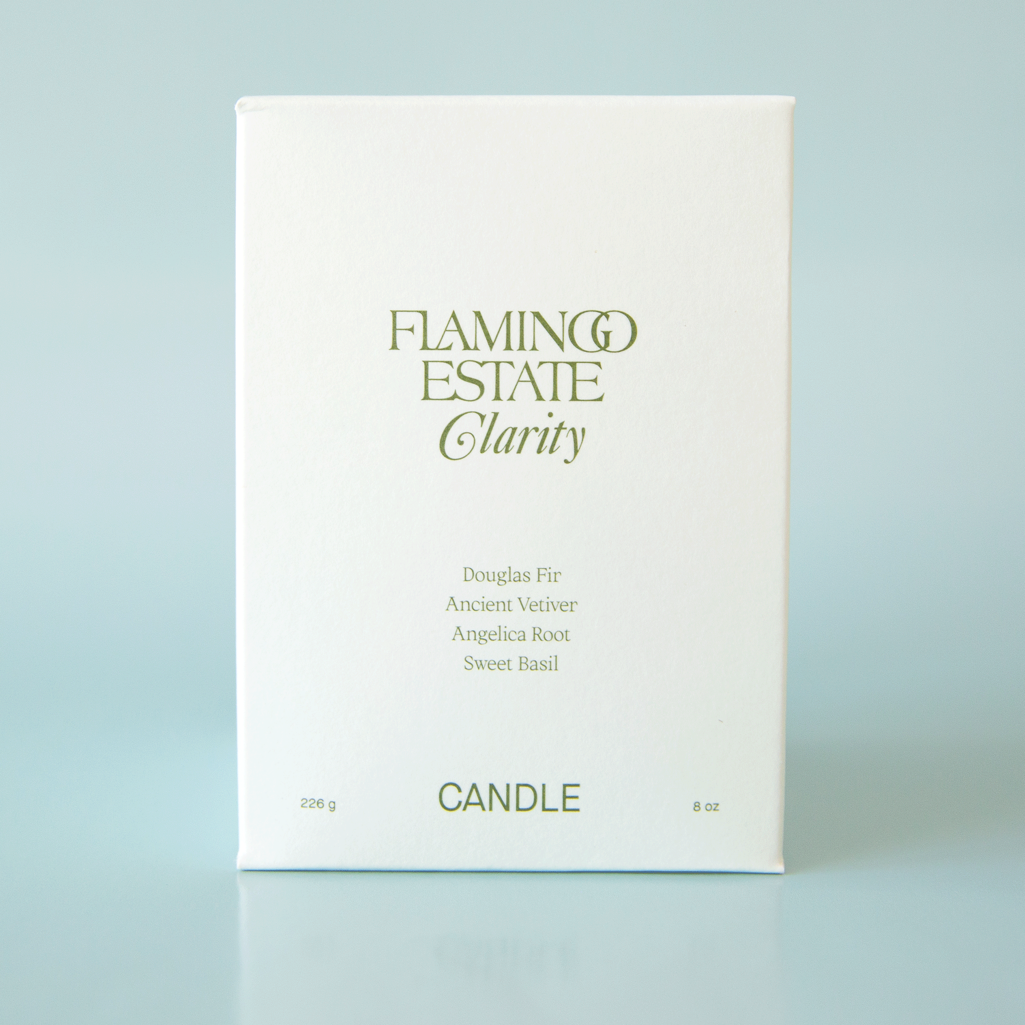 On a blue background is a box containing a green glass candle. The front of the box has green text that reads, "Flamingo Estate Clarity Douglas Fir Ancient Vetiver Angelica Root Sweet Basil".