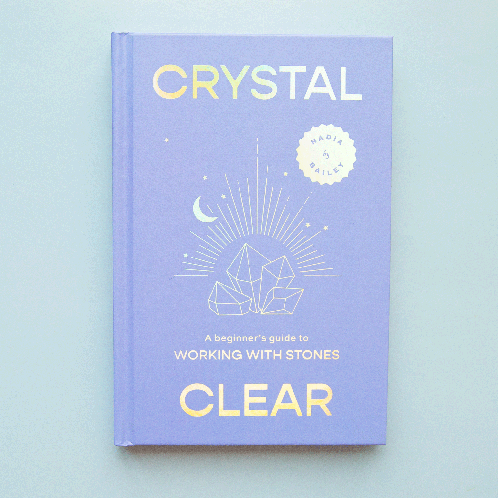 On a blue background is a periwinkle book cover with iridescent text on the front that reads, &quot;Crystal Clear A beginners guide to Working With Stones&quot;. 