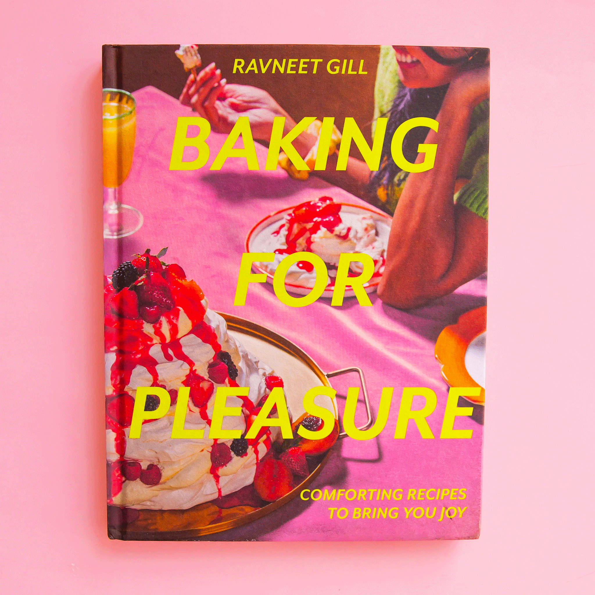 On a pink background is a hot pink book cover with desserts on the front and yellow text that reads, &quot;Baking For Pleasure&quot;. 