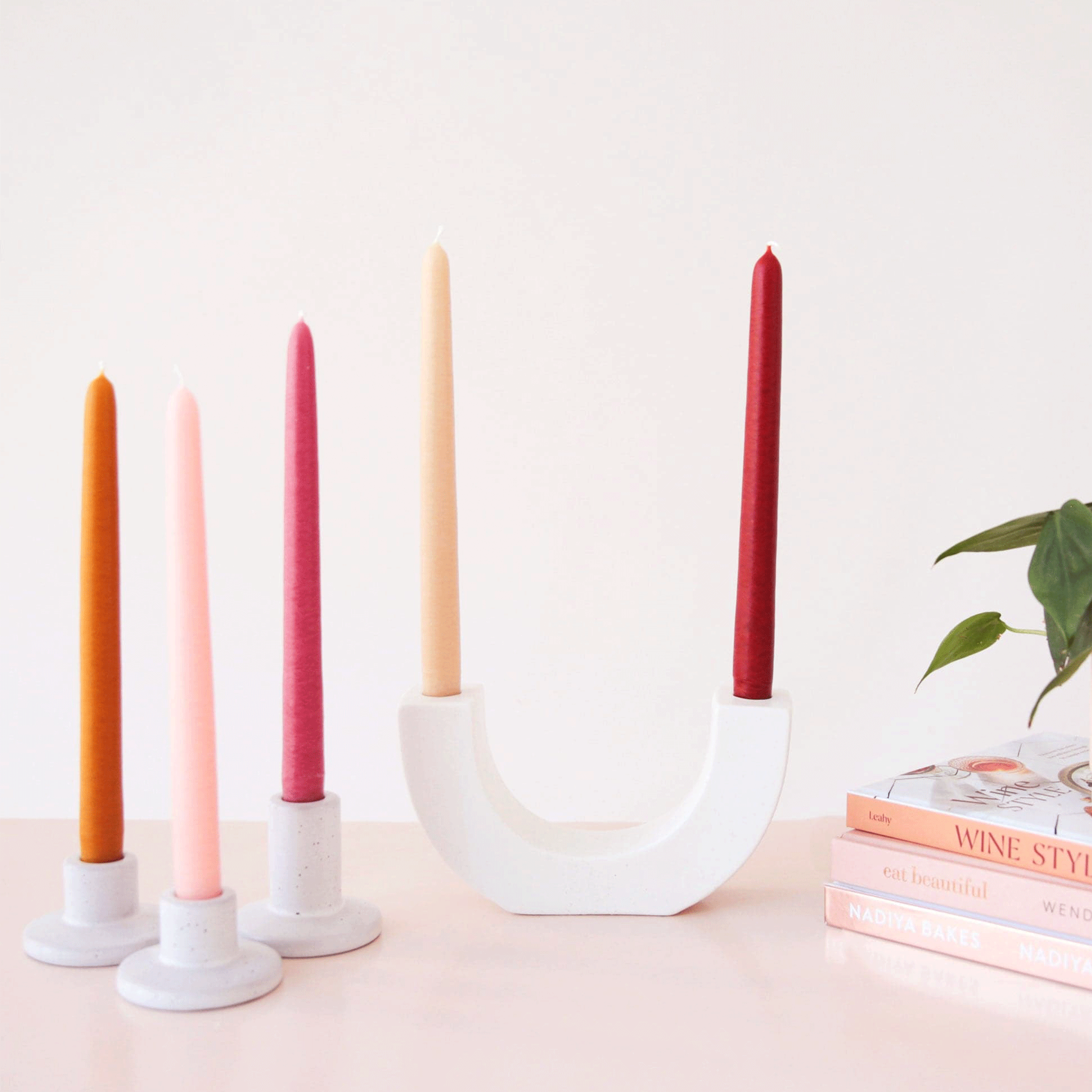 A set of five taper candles in warm shades of pink, orange and reds along photographed here in a U-shaped taper candle holder and individual ceramic ones on a table scape with books and a plant.