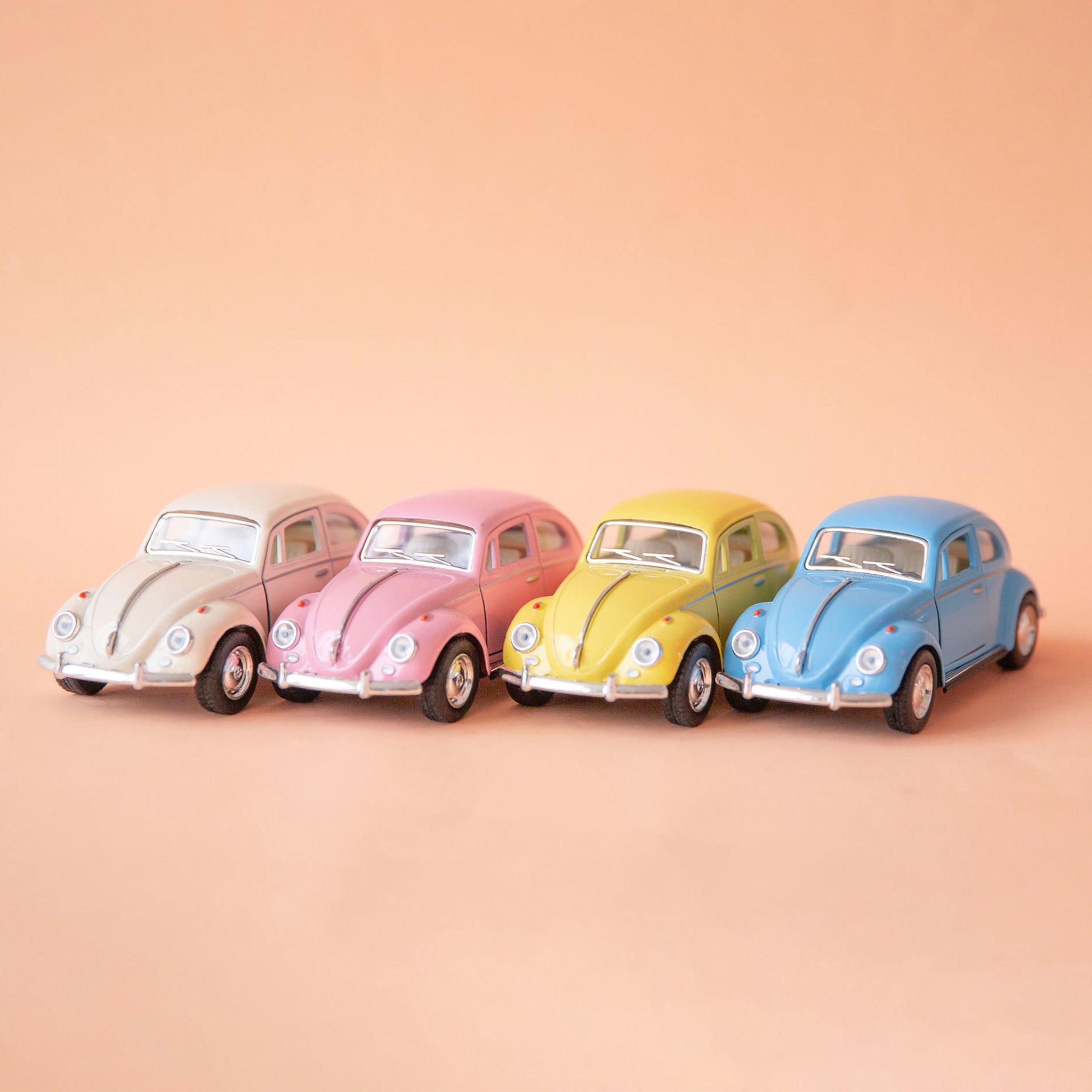 On a tan background is all four toy Volkswagen Beetle cars in a white, pink, yellow and blue color. 