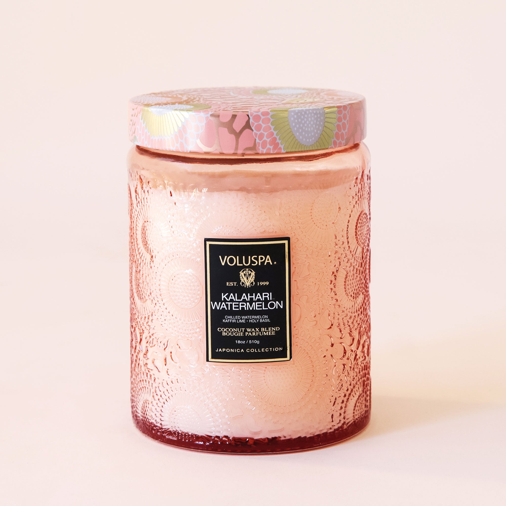Pink colored embossed candle with patterned colorful lid. The embossed pattern and lid have an abstract floral design with use of the colors peach, gold, and white.