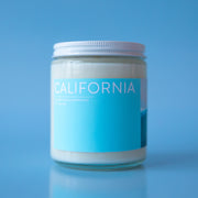 On a blue background is a clear glass jar candle with a light blue rectangular label across the front the reads, "California" in white letters and a white screw on lid.