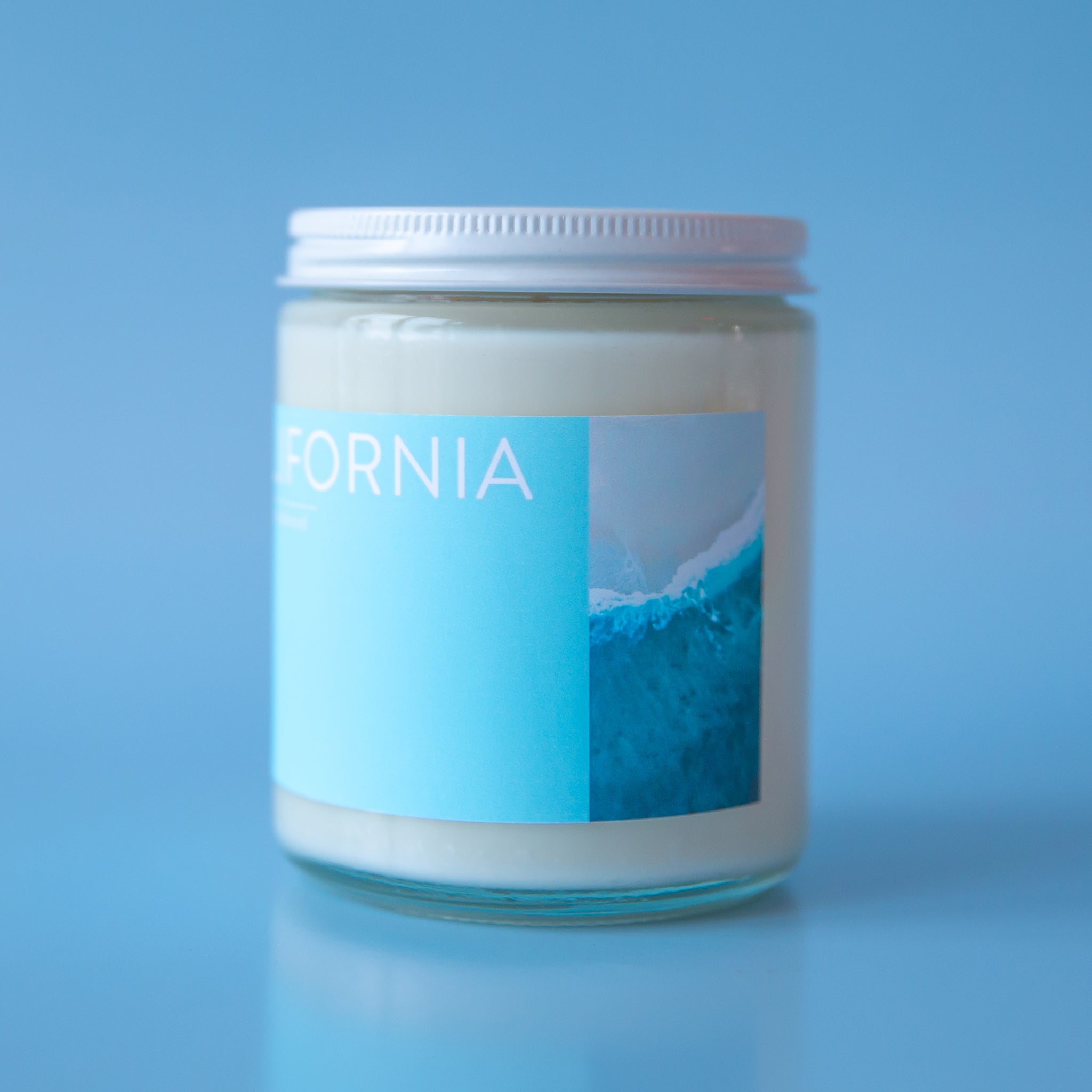On a blue background is a clear glass jar candle with a light blue rectangular label across the front the reads, &quot;California&quot; in white letters and a white screw on lid.