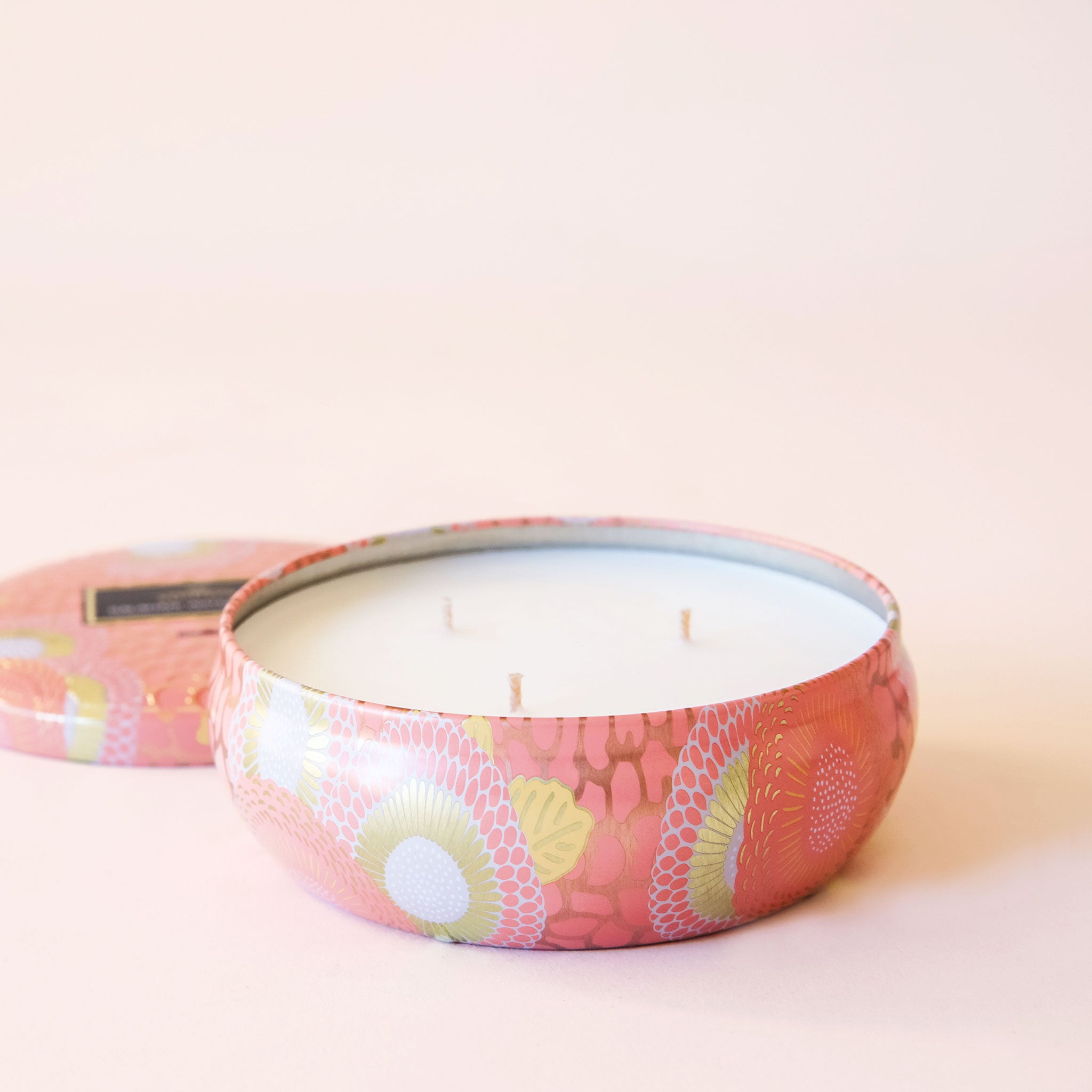 A round tin 3 wick candle with an abstract floral and spotted design. The metal from the tin reflects light in a beautiful way, especially on its gold parts. The other colors on this tin are white and and a peach color.